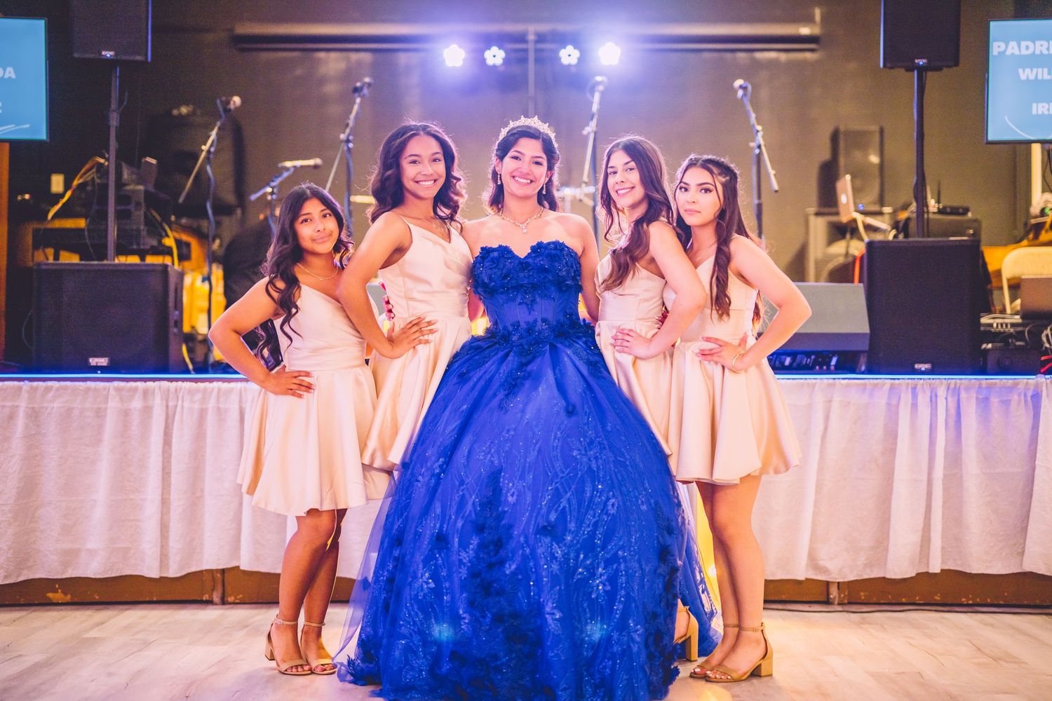 Group celebrating at a quinceañera or formal event with person in royal blue ball gown centerpiece on dance floor.