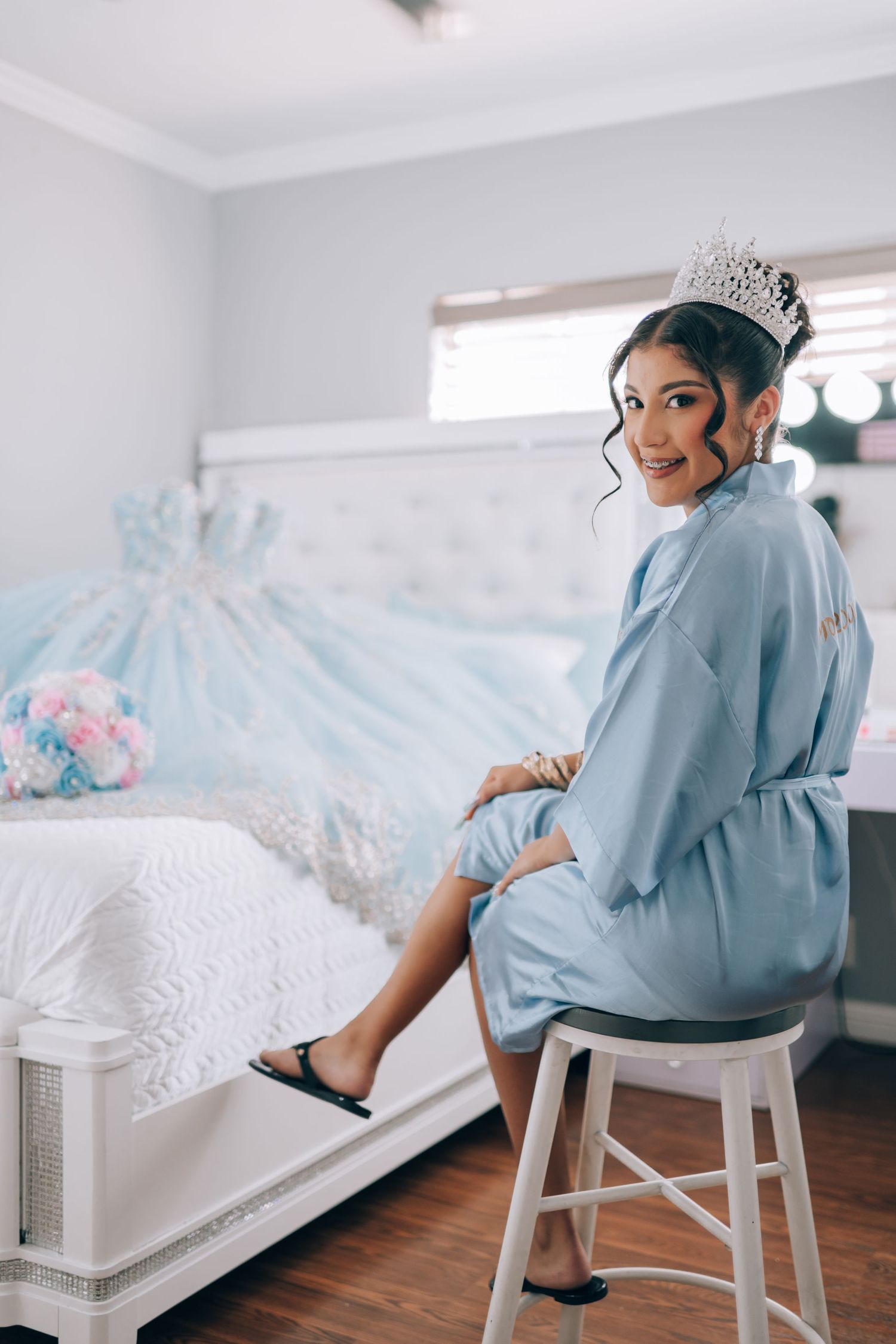 Elegant figure in light blue robe and tiara sits on white stool beside bed with pale blue ballgown displayed.