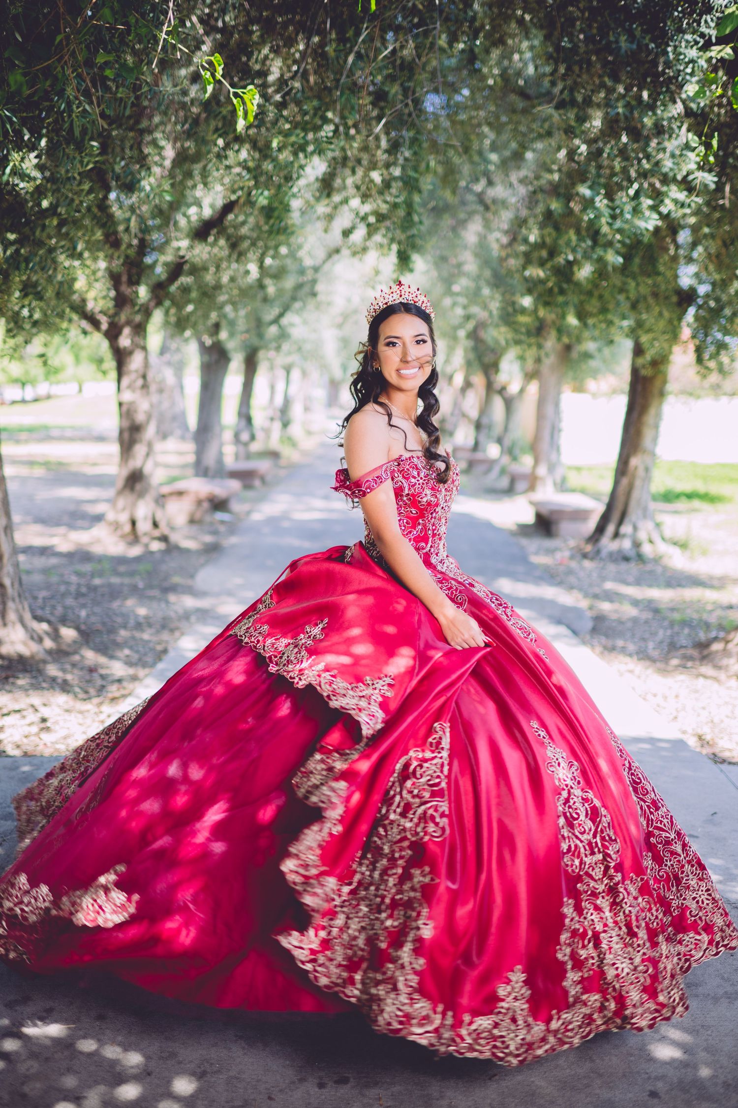 A stunning red quinceañera ball gown with intricate gold embroidery photographed in a tree-lined pathway.