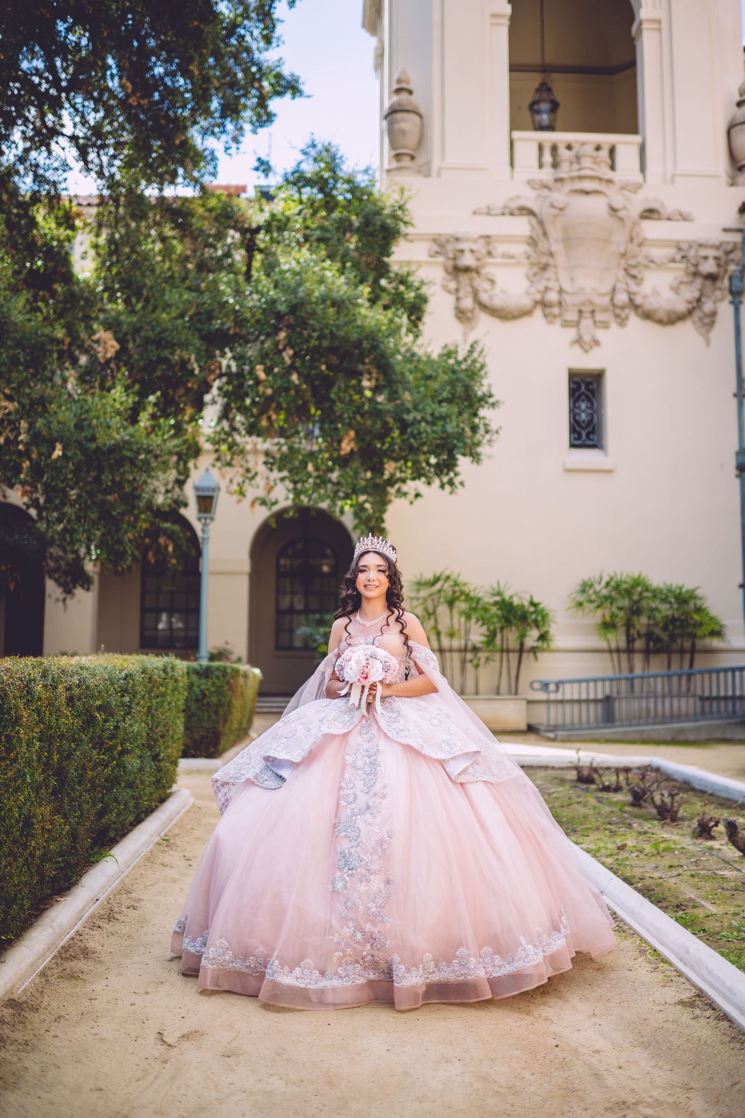 Young woman in pink quinceañera ball gown with lace trim and tiara poses outdoors near elegant building.