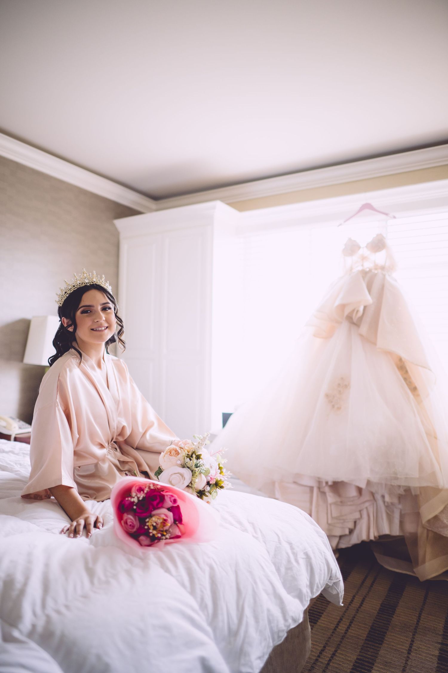 A bride in a silk robe sits on a bed with flowers while her white wedding gown hangs in the background.