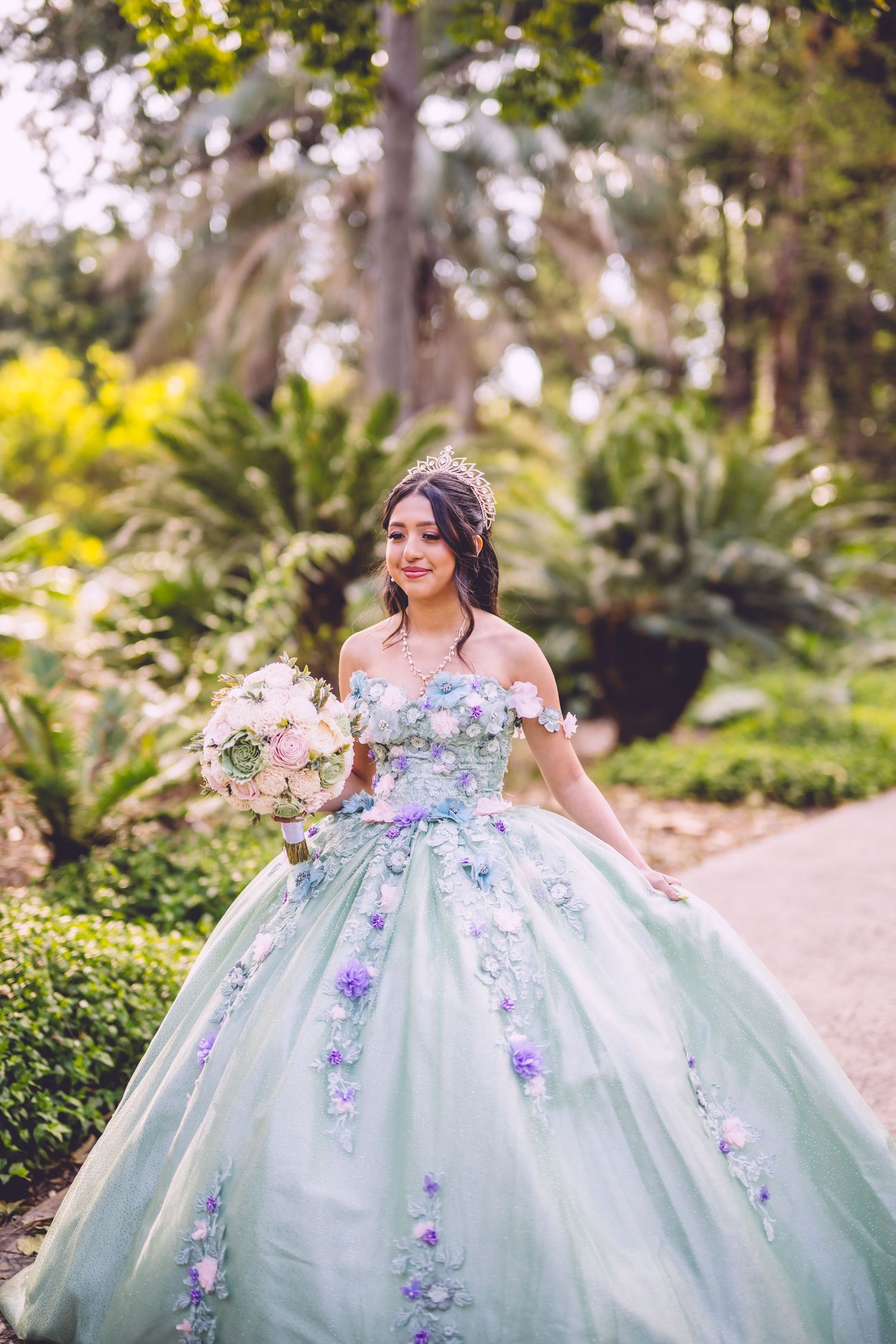 A person in a mint green ballgown with floral appliques holds a pastel bouquet while posing in a lush garden setting.