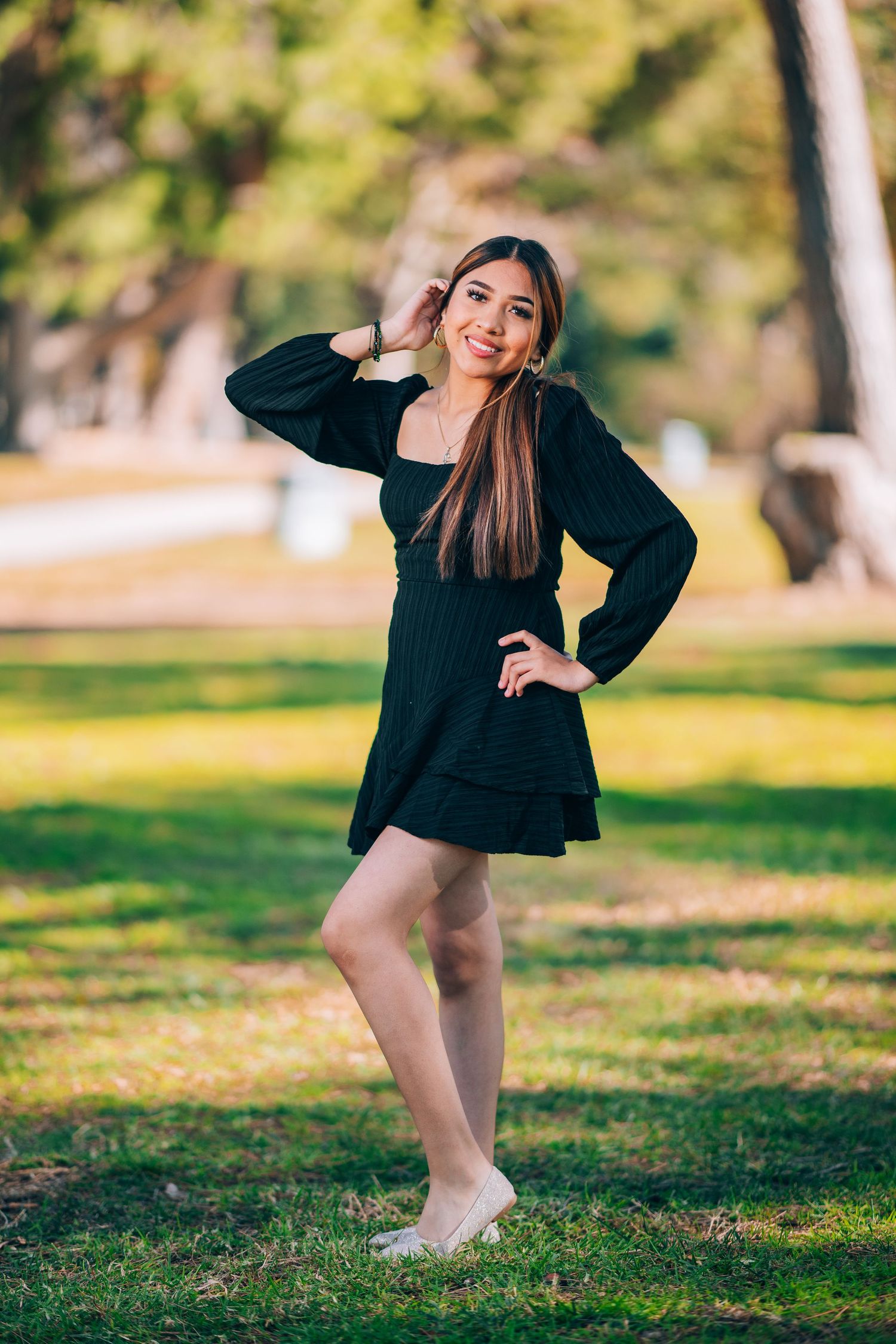 Young woman in black mini dress posing outdoors in a sunny park, smiling with long brown hair.