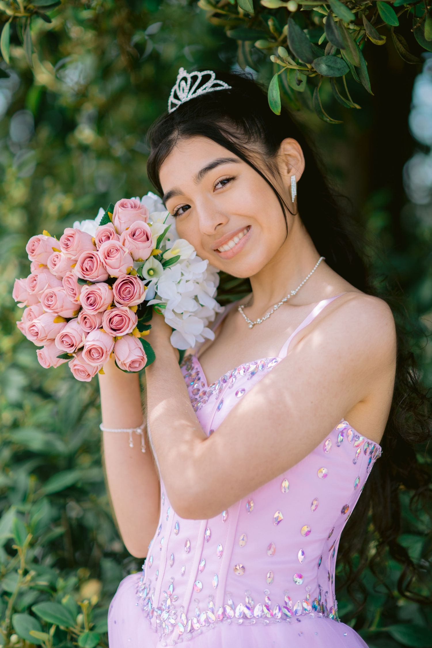 A person in a pink dress poses with pink roses and a tiara in a garden setting with dappled sunlight.