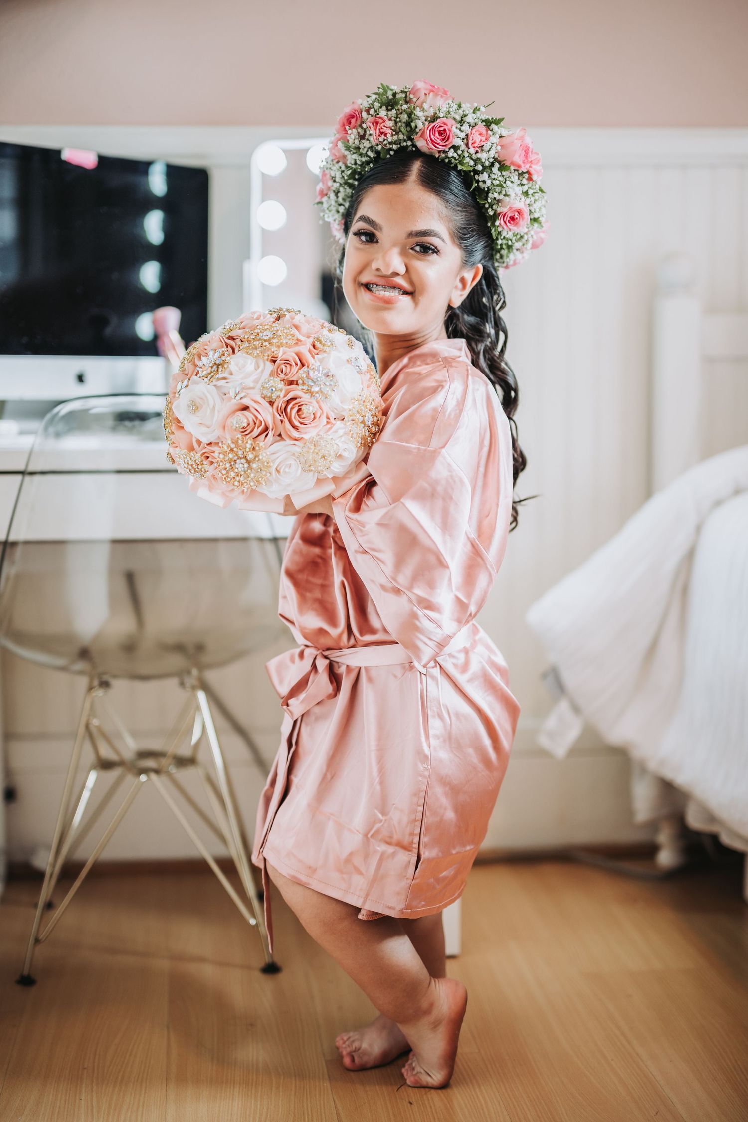 A flower crown adorned figure in pink satin robe holds peachy rose bouquet in bright room.