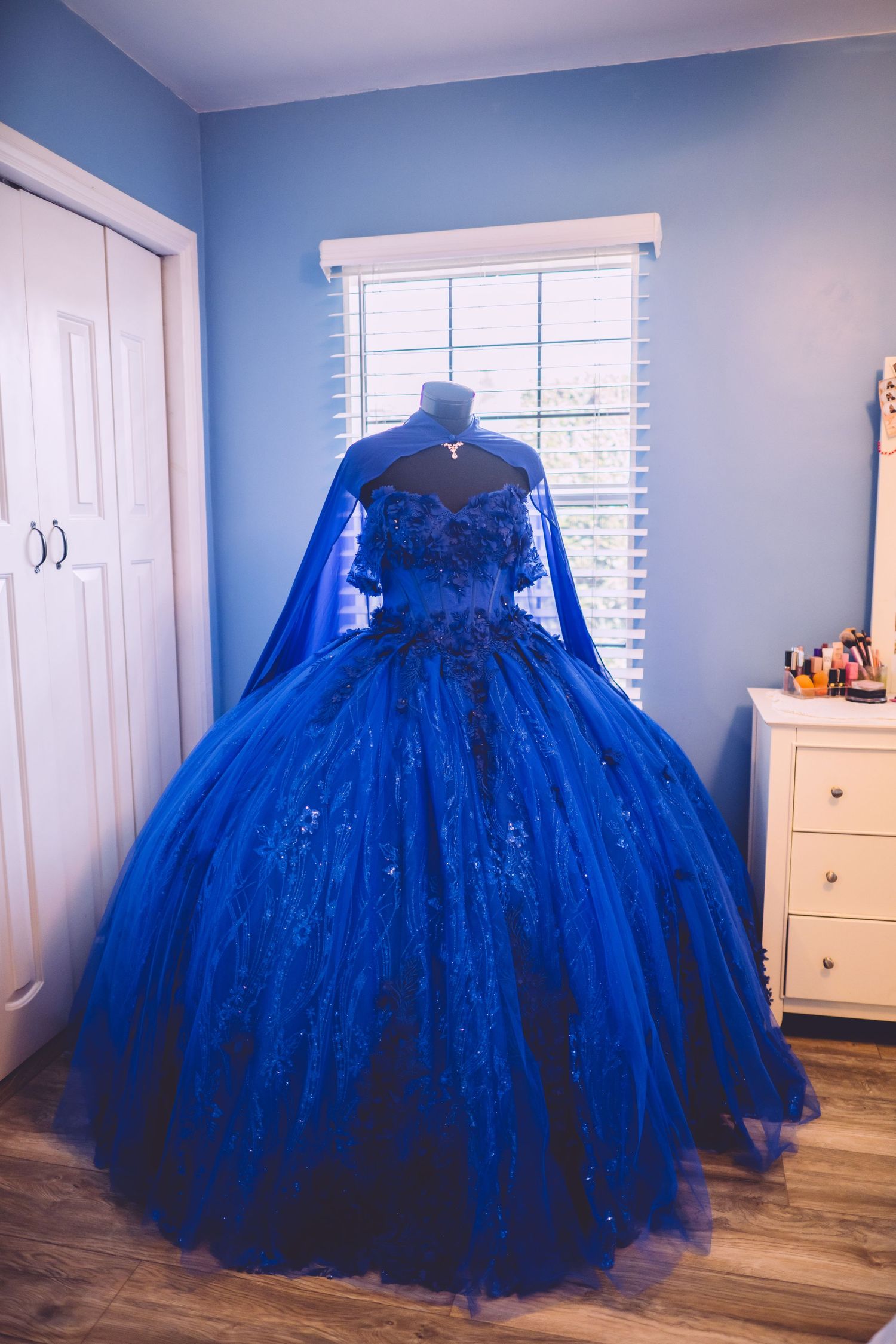 A stunning royal blue ballgown with sparkles and a flowing cape displayed against light blue walls in a bedroom.