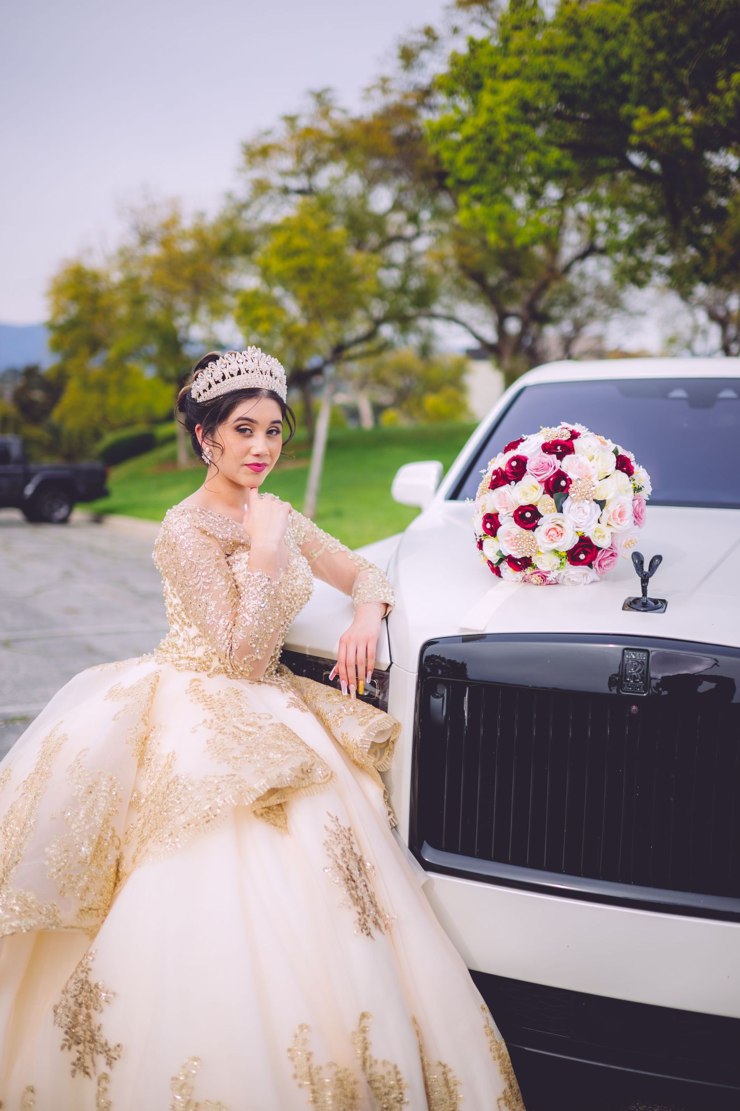 A luxurious cream and gold ballgown with a tiara is showcased next to a white Rolls Royce with a red and white bouquet.