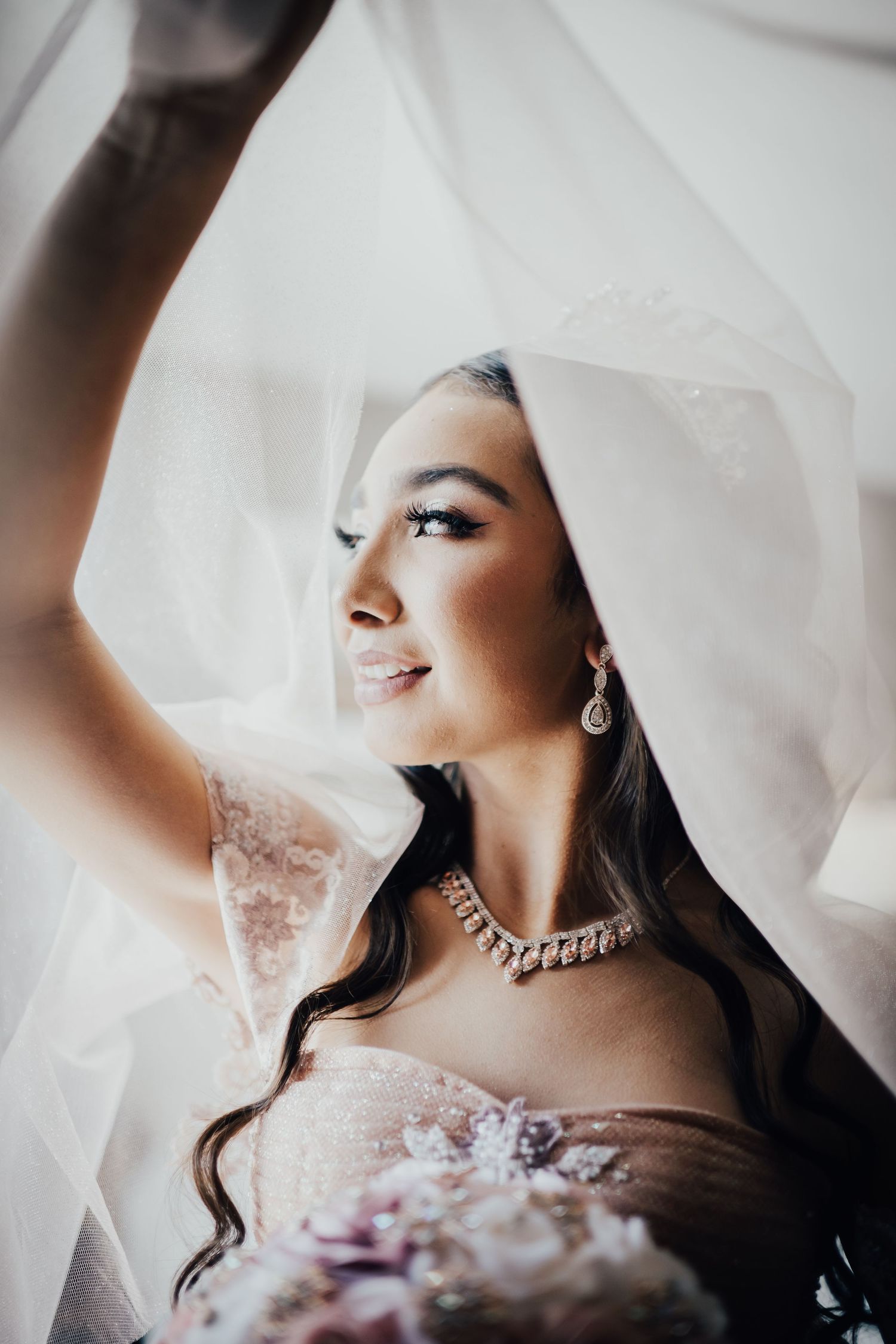 Elegant bride in lace gown holding bouquet under veil, wearing diamond necklace and earrings, gazing softly to the side.