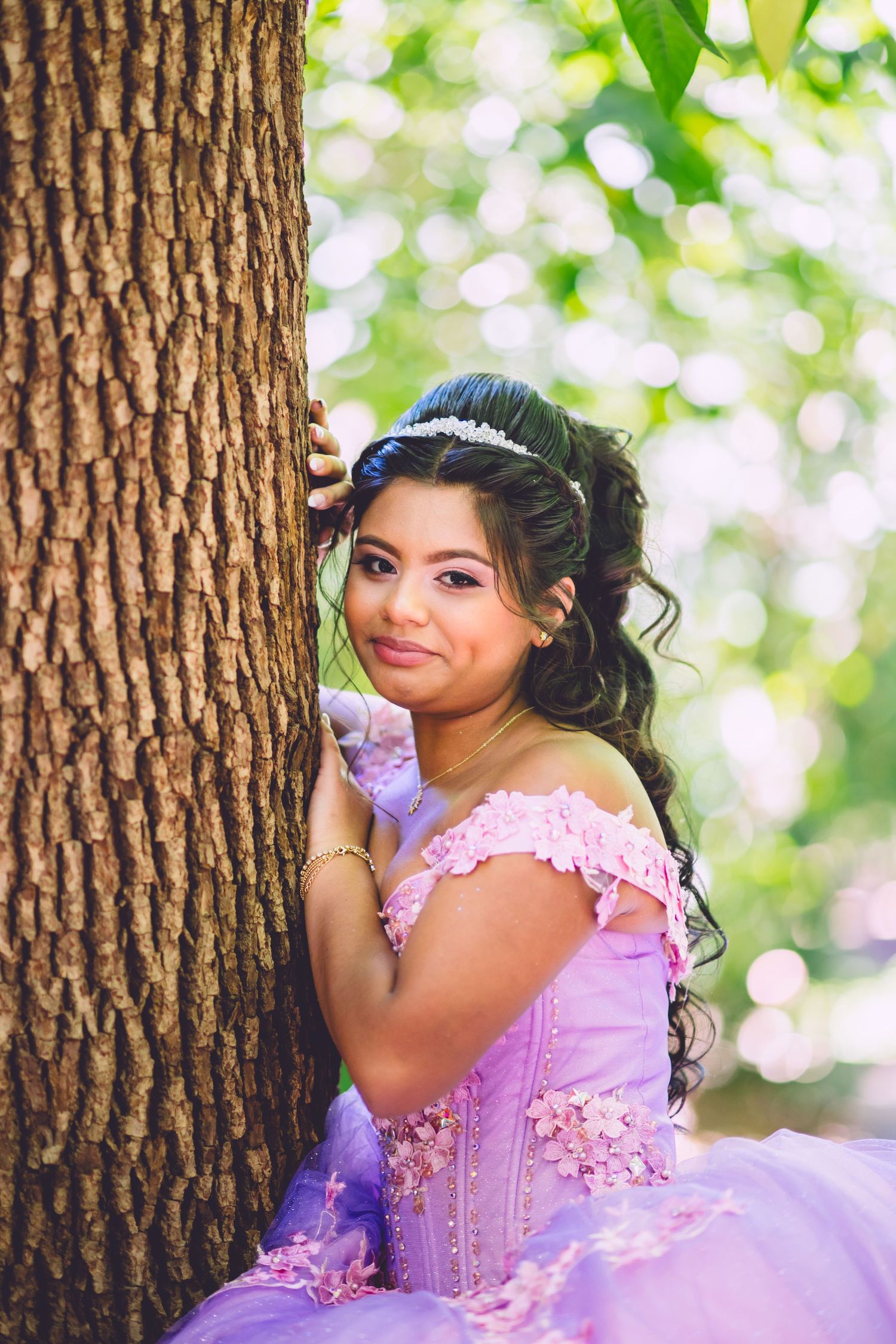 A woman in a lavender quinceañera dress poses next to a tree trunk with a dreamy bokeh background.
