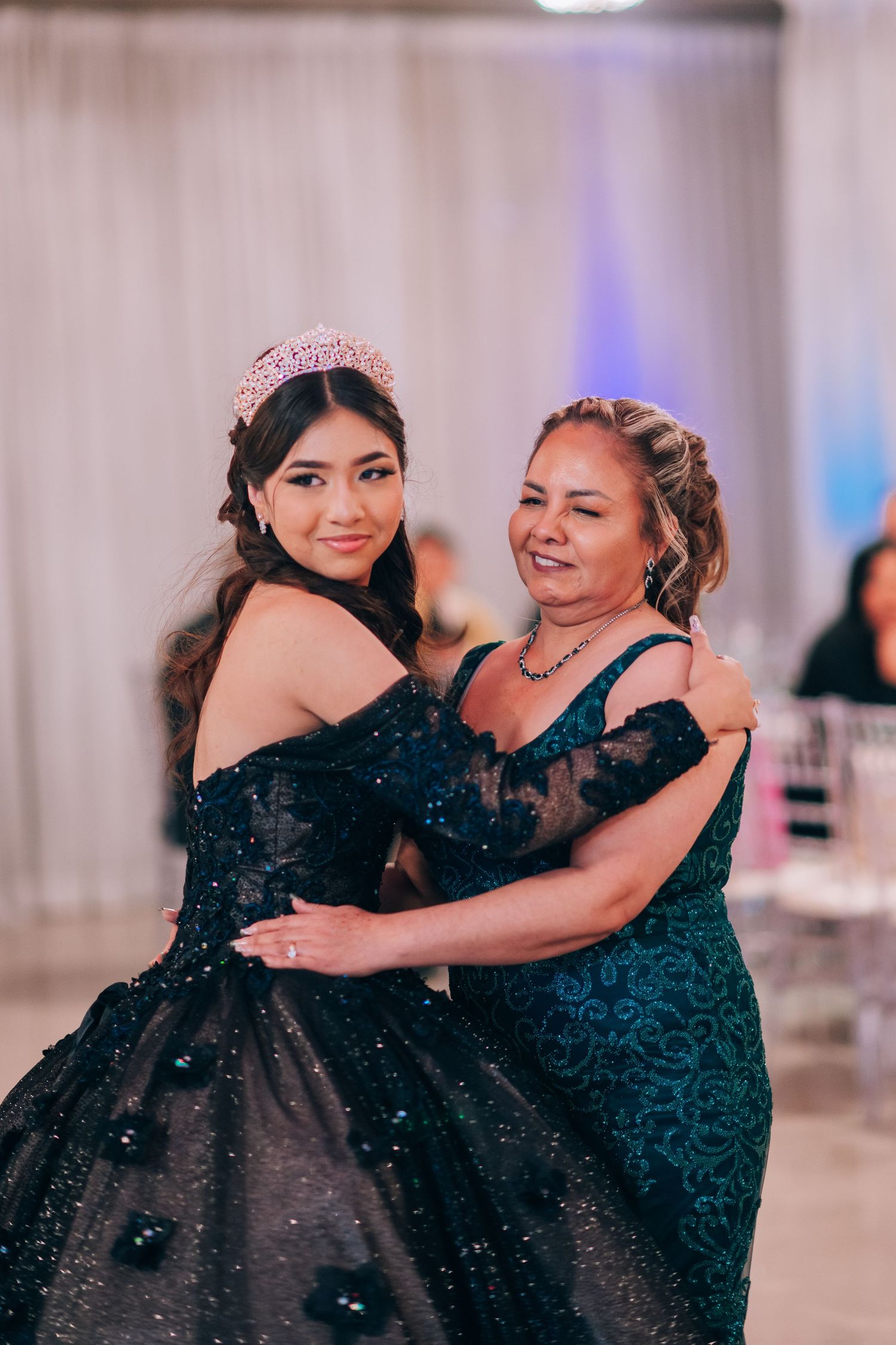 Two women in sparkling black formal gowns embrace while posing at an elegant event with decorative lighting.