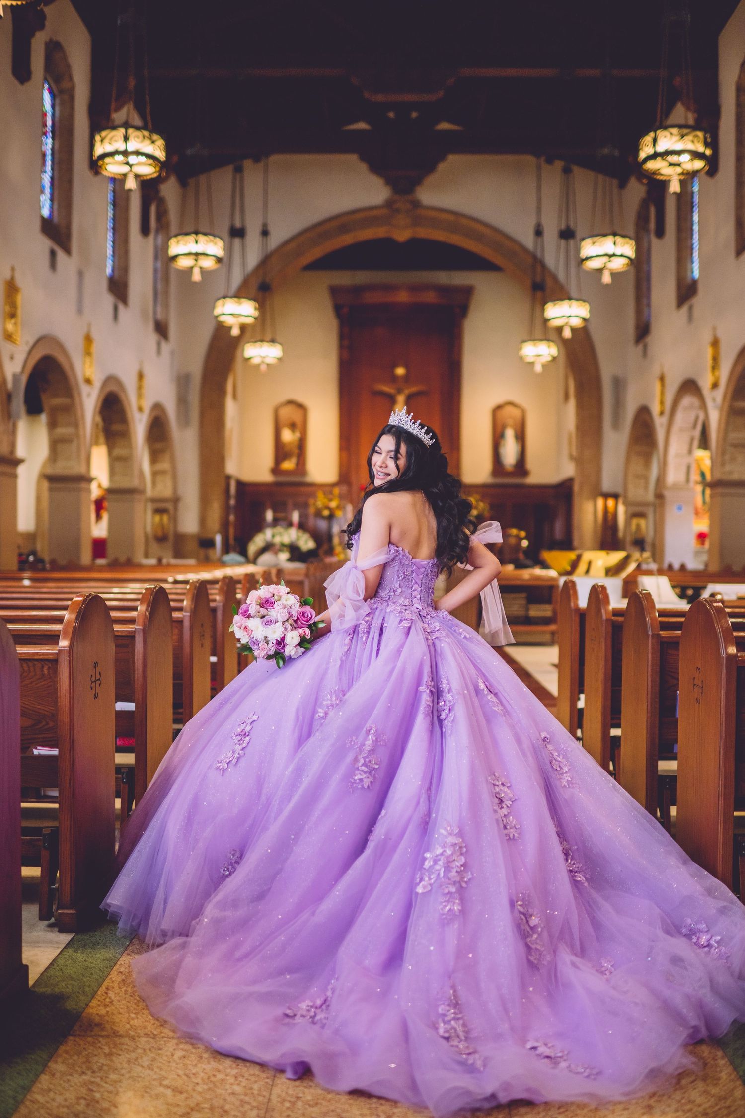 Young woman in lavender quinceañera ball gown with floral appliqués and tiara poses in ornate church aisle.