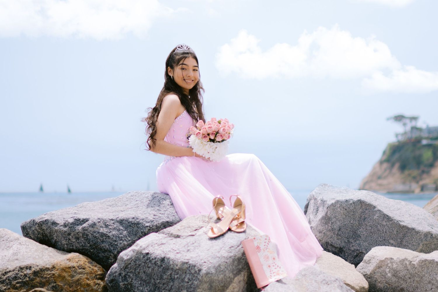 Young woman in pink gown holding bouquet sits on rocks by ocean wearing tiara and heels nearby.
