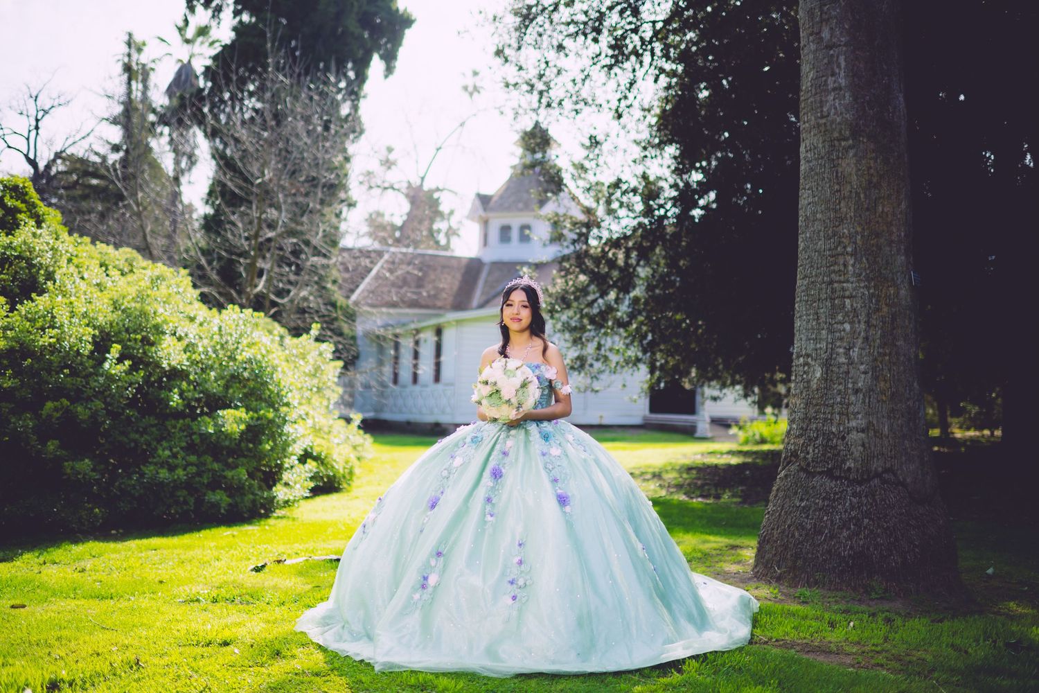 Young woman in a light blue ball gown holding a bouquet, posing in a lush garden near a white chapel.