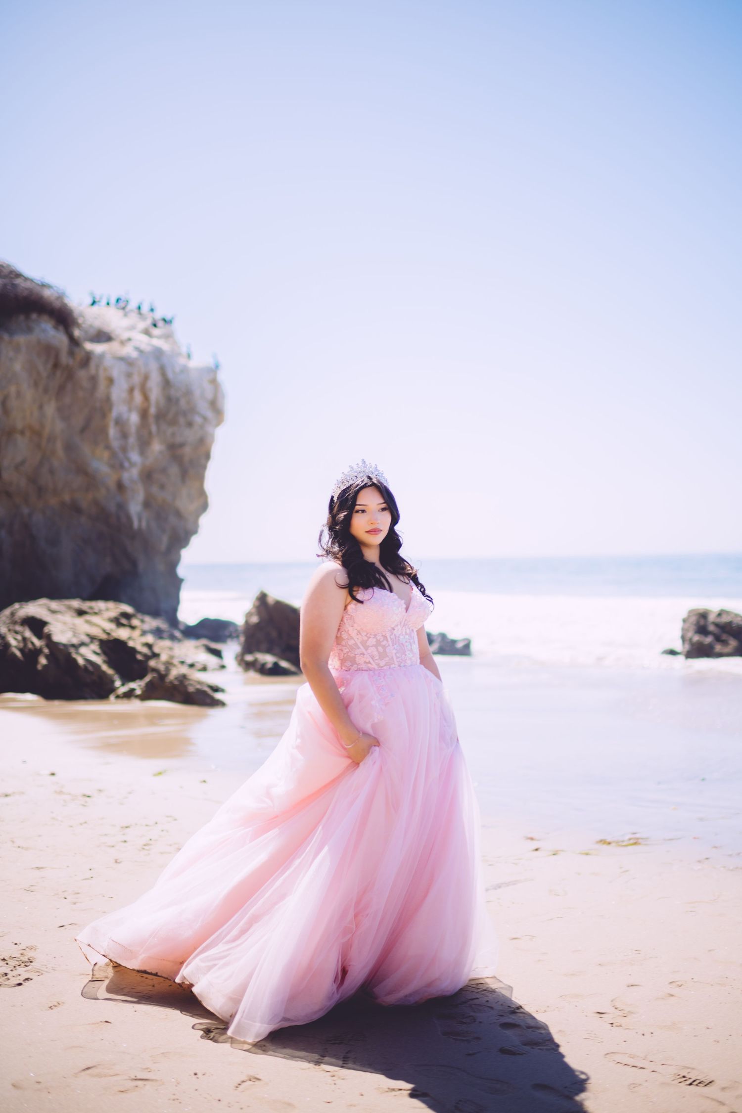 A person in a flowing pink gown poses gracefully on a beach with rocky cliffs in the background.