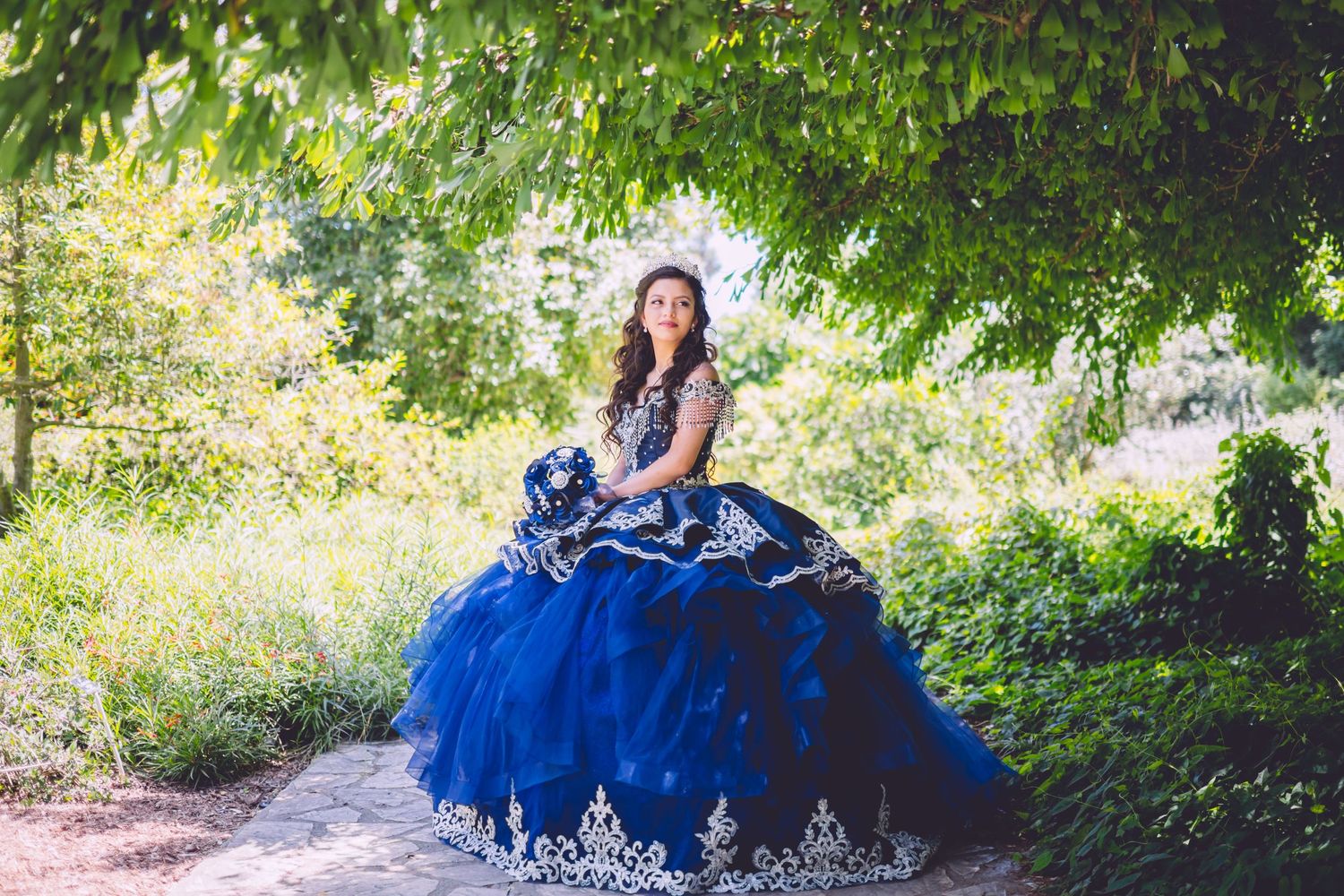 A figure in a flowing royal blue ball gown poses elegantly in a garden setting with lush greenery.