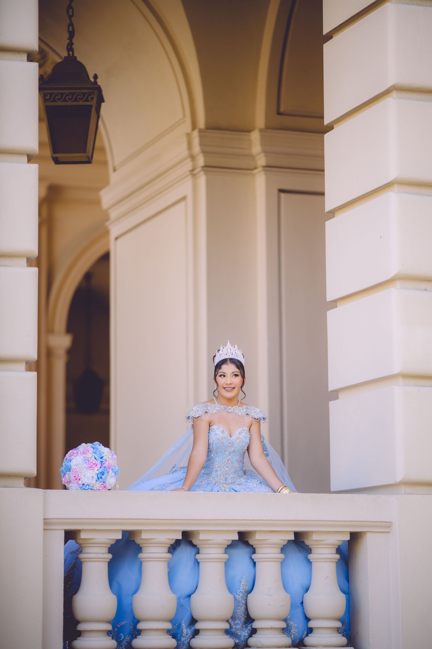 A bride in a blue and white gown poses elegantly on a balcony with classical architectural columns.