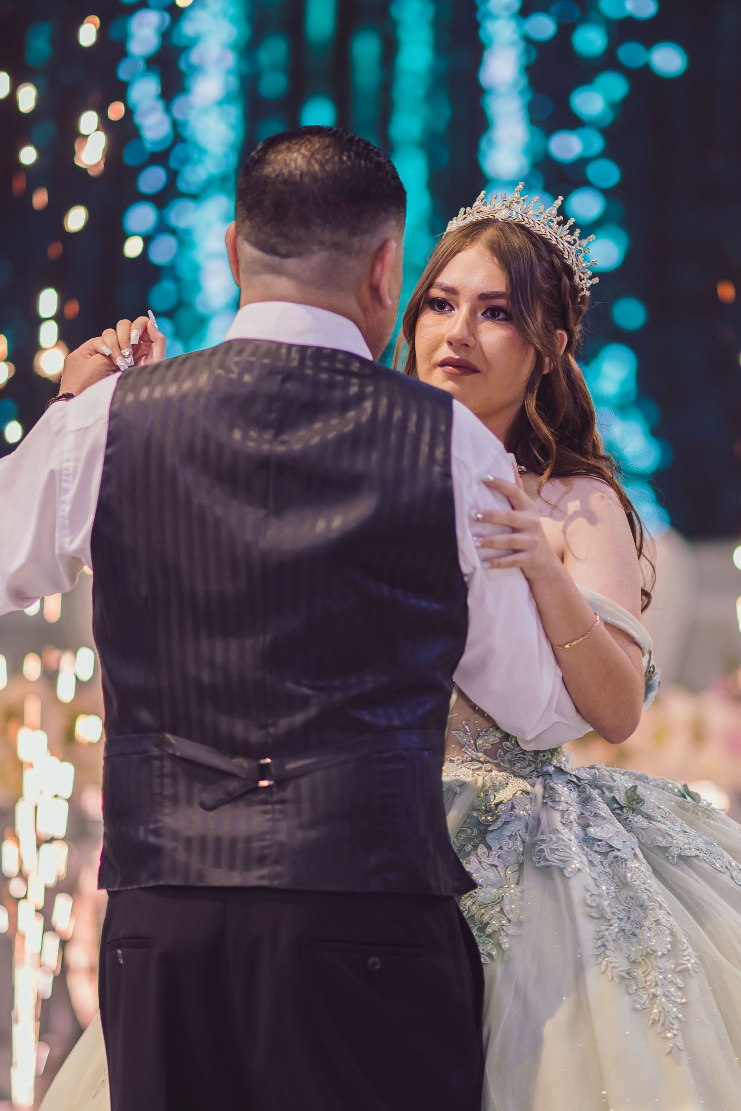 Bride in crown and lace gown shares first dance with groom in black vest at elegant wedding reception.