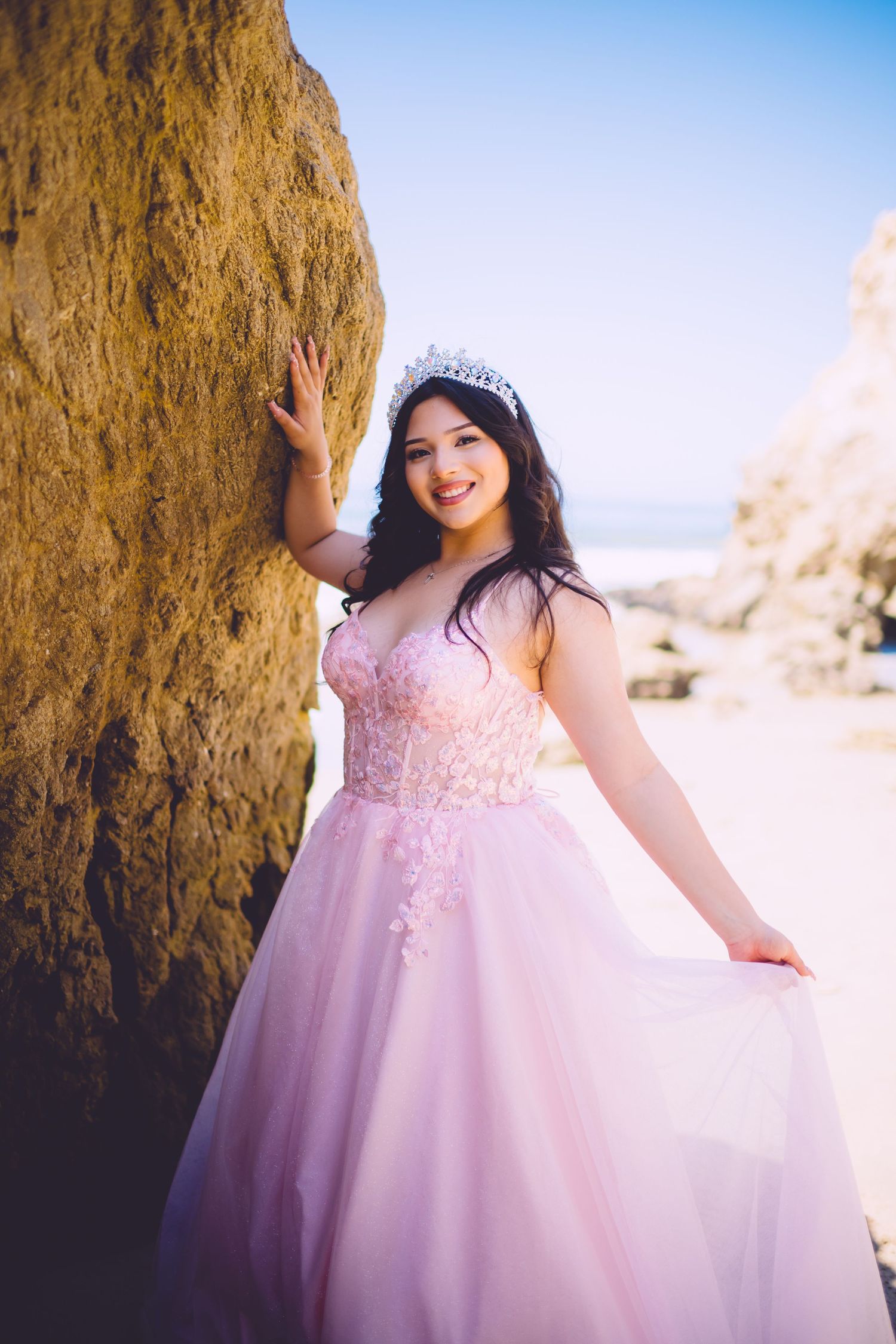 A person in a flowing pink princess gown and tiara poses against a rocky beach backdrop on a sunny day.