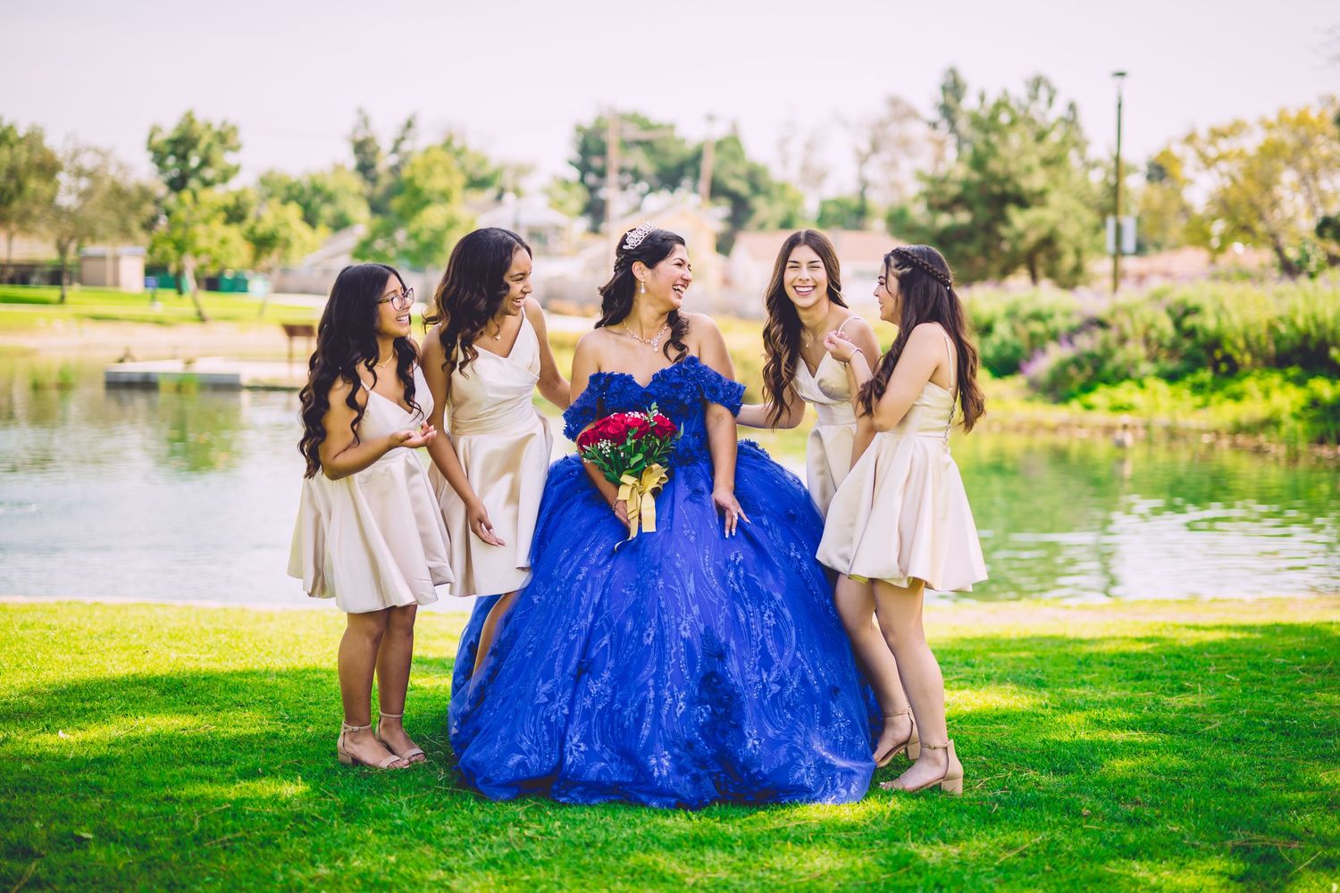 A young woman in a royal blue quinceañera gown holds red roses, surrounded by four bridesmaids in white dresses by a pond.