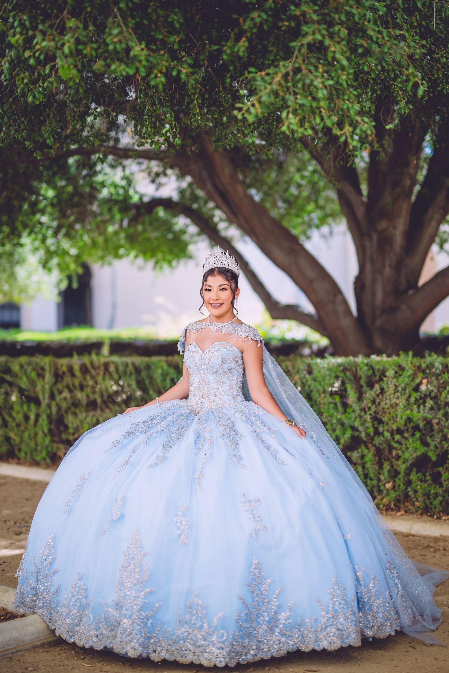 A young girl in a light blue quinceañera ball gown with lace details poses outdoors under trees for a photoshoot.