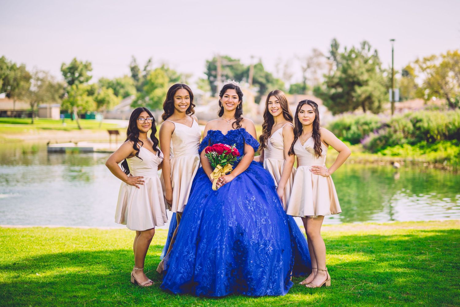 A group in formal attire poses on grass near a lake, with center figure wearing a vibrant royal blue ballgown.