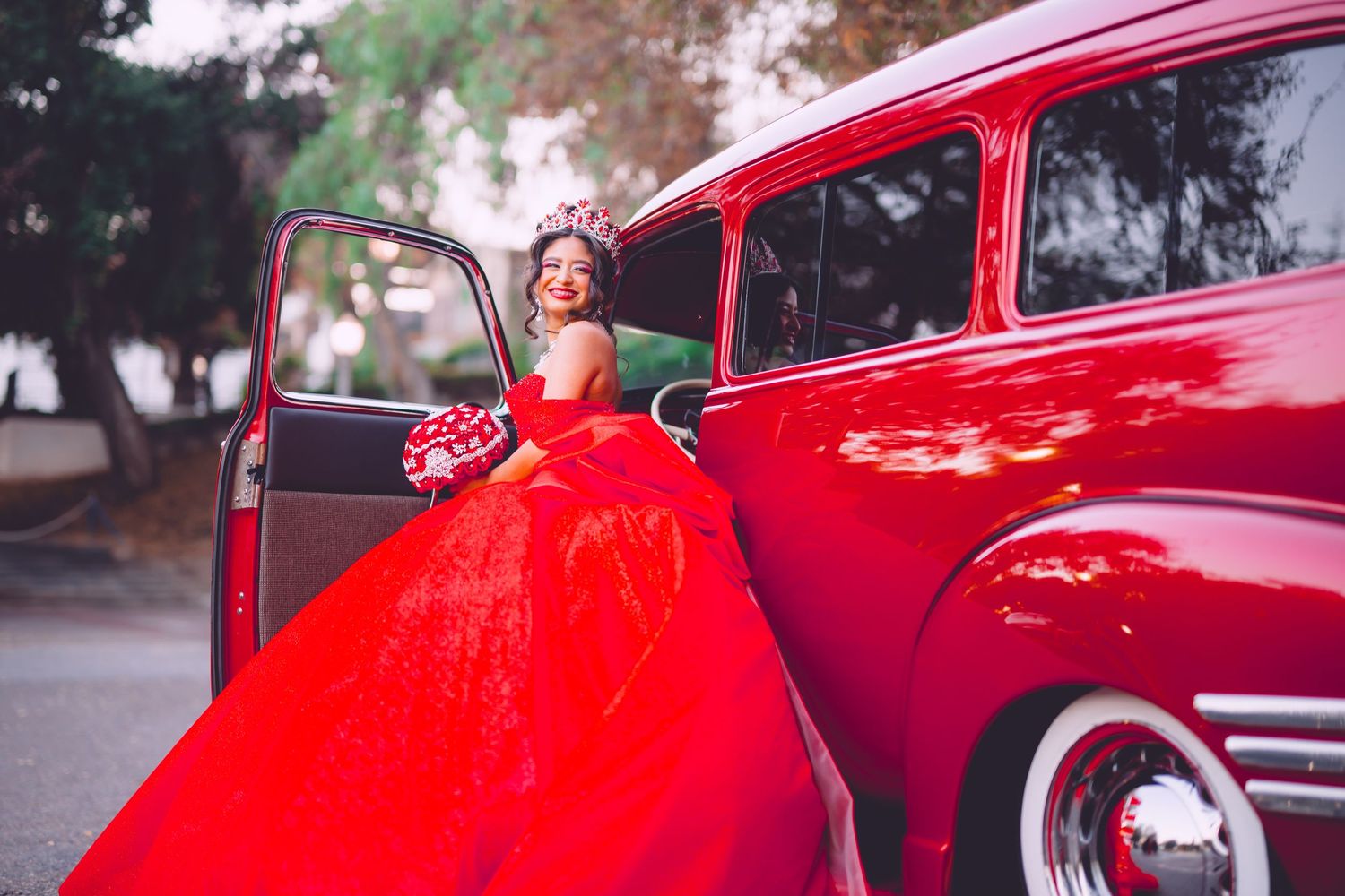 A red ballgown sweeps dramatically beside a vintage red classic car parked on a street.