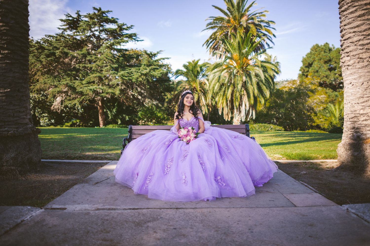 A person in a flowing lavender ballgown poses in a garden setting with palm trees and lush greenery.