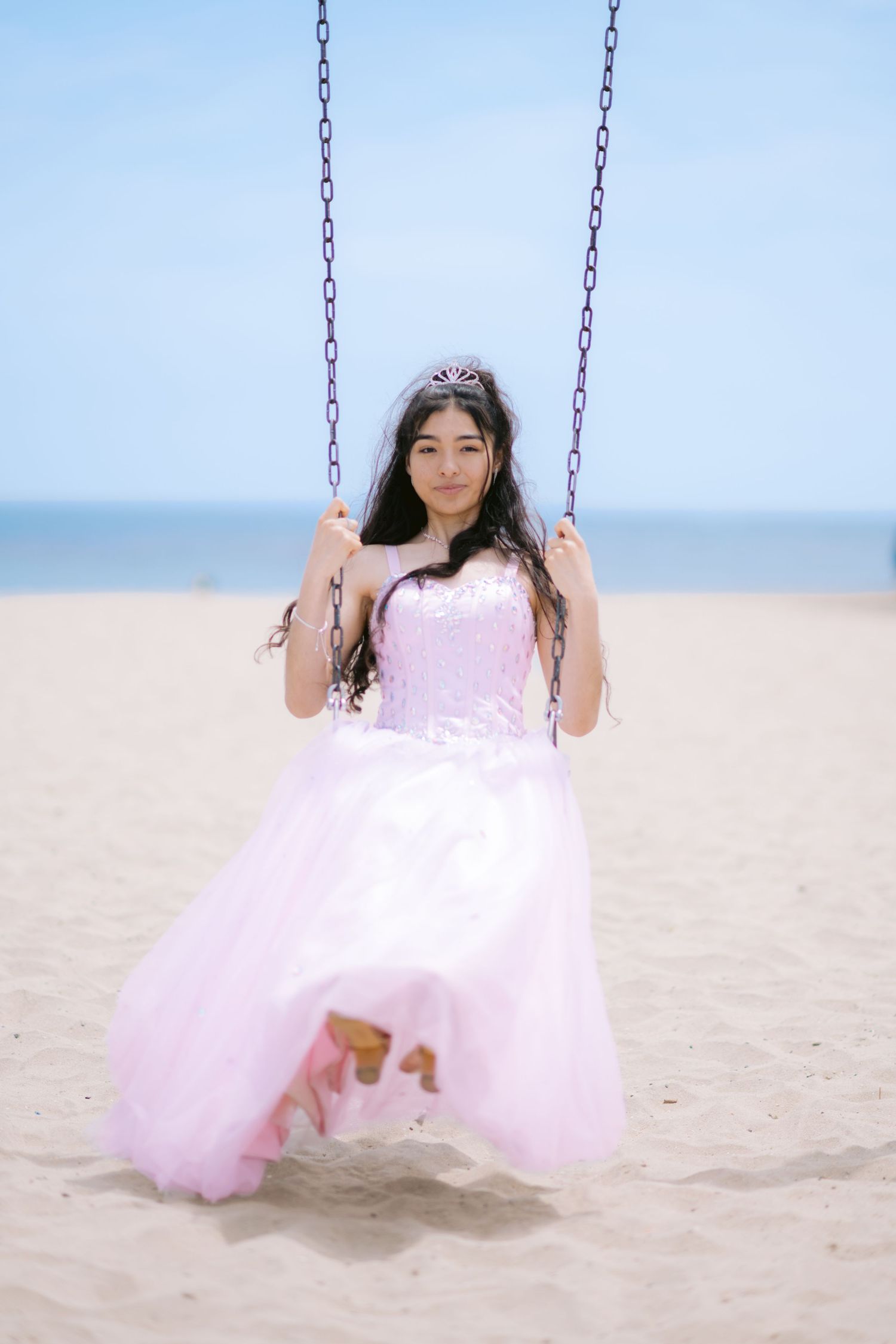 A person in a flowing pink tulle gown sits gracefully on a swing at the beach during a photoshoot.