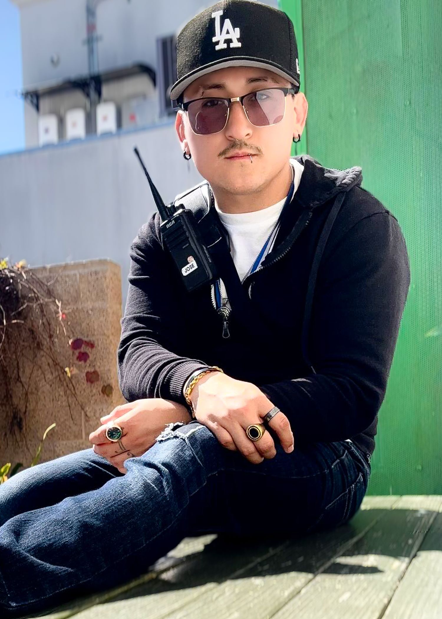 A casual portrait of someone wearing an LA Dodgers cap, sunglasses and black jacket while sitting against a green wall.