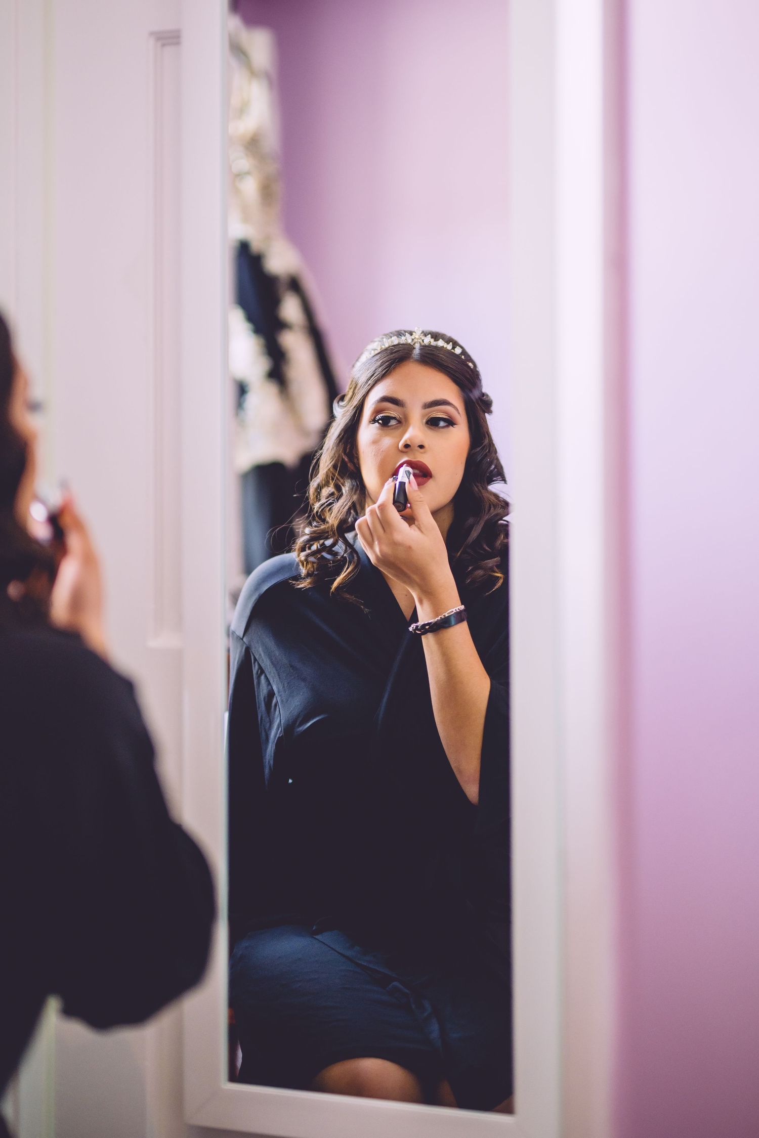 Mirror reflection shows someone in navy dress wearing a tiara while applying makeup in pink-tinted room.