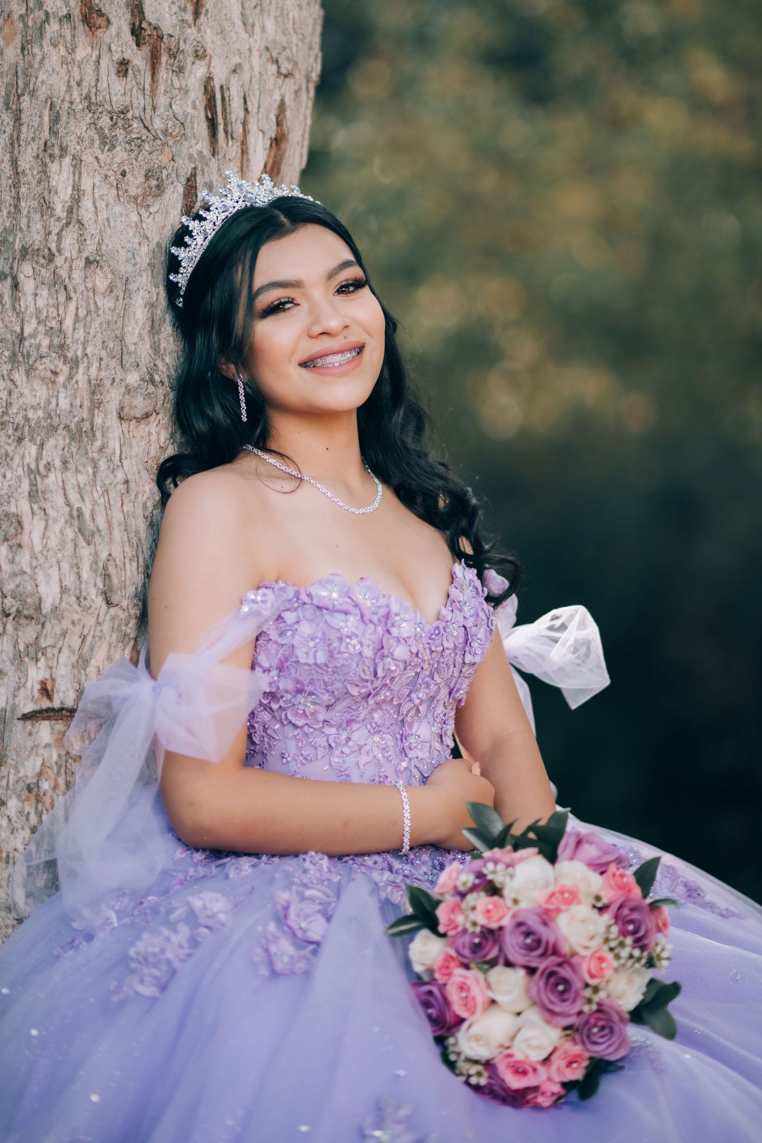 Young woman in lavender quinceañera dress with tiara poses by tree holding pink and purple floral bouquet.