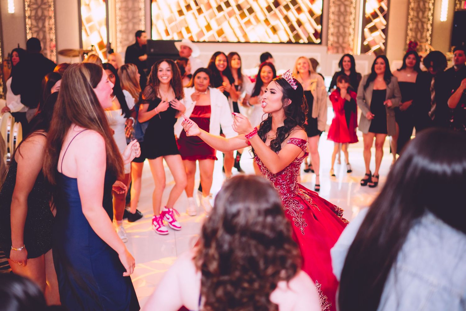 A lively group of people dancing and celebrating at a party with decorative lighting in the background.