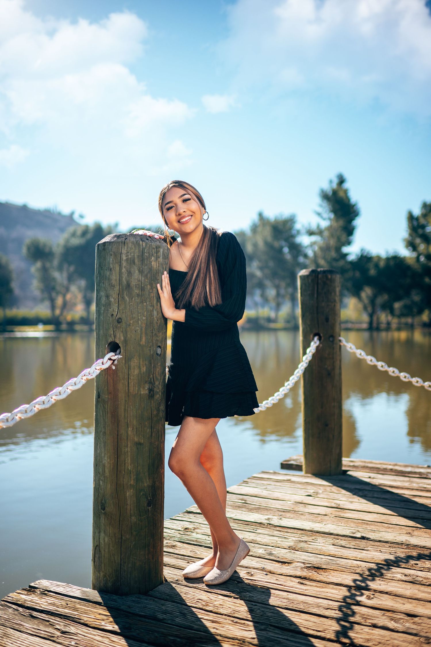 Young woman in black dress posing on wooden dock by lake, smiling beside rope chain and post.