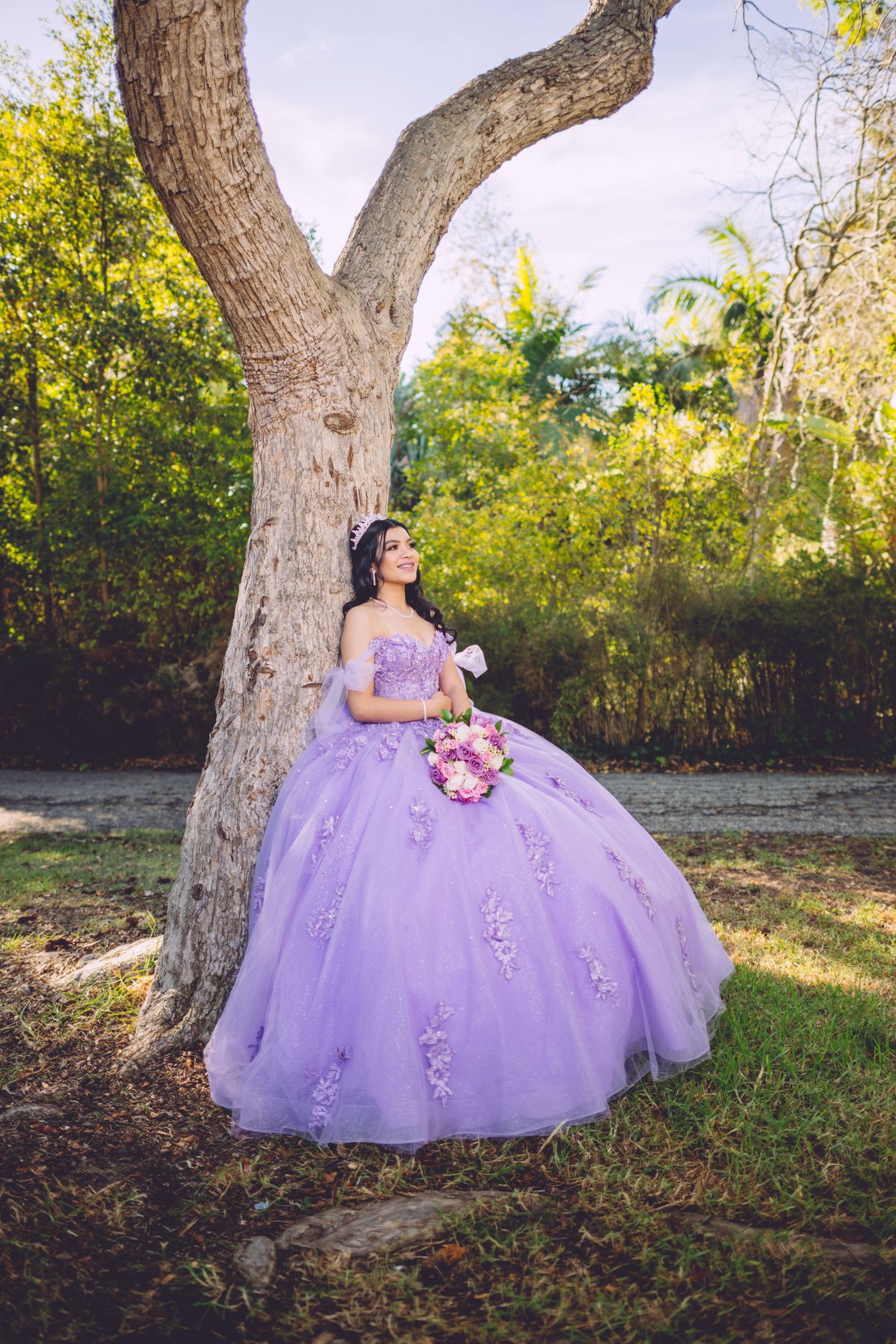 A person in a lavender quinceanera ball gown poses elegantly under a large tree in an outdoor garden setting.
