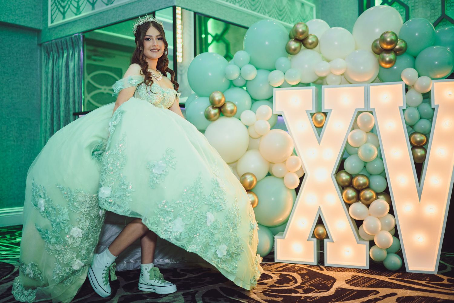 Young woman in a mint green quinceañera ball gown poses in front of XV marquee letters and balloon backdrop.