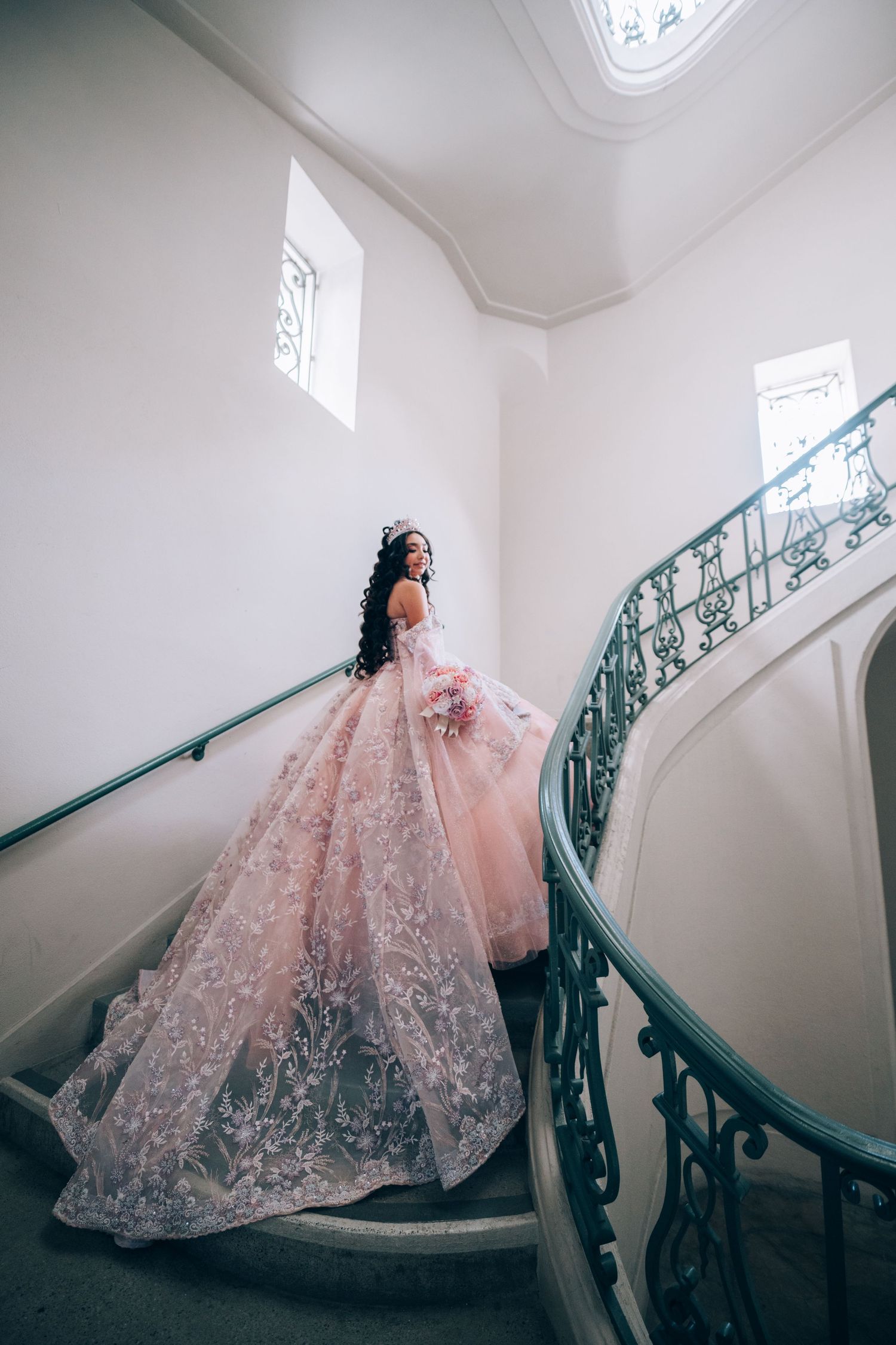 Ethereal pink ball gown with lace details on grand spiral staircase in bright interior.