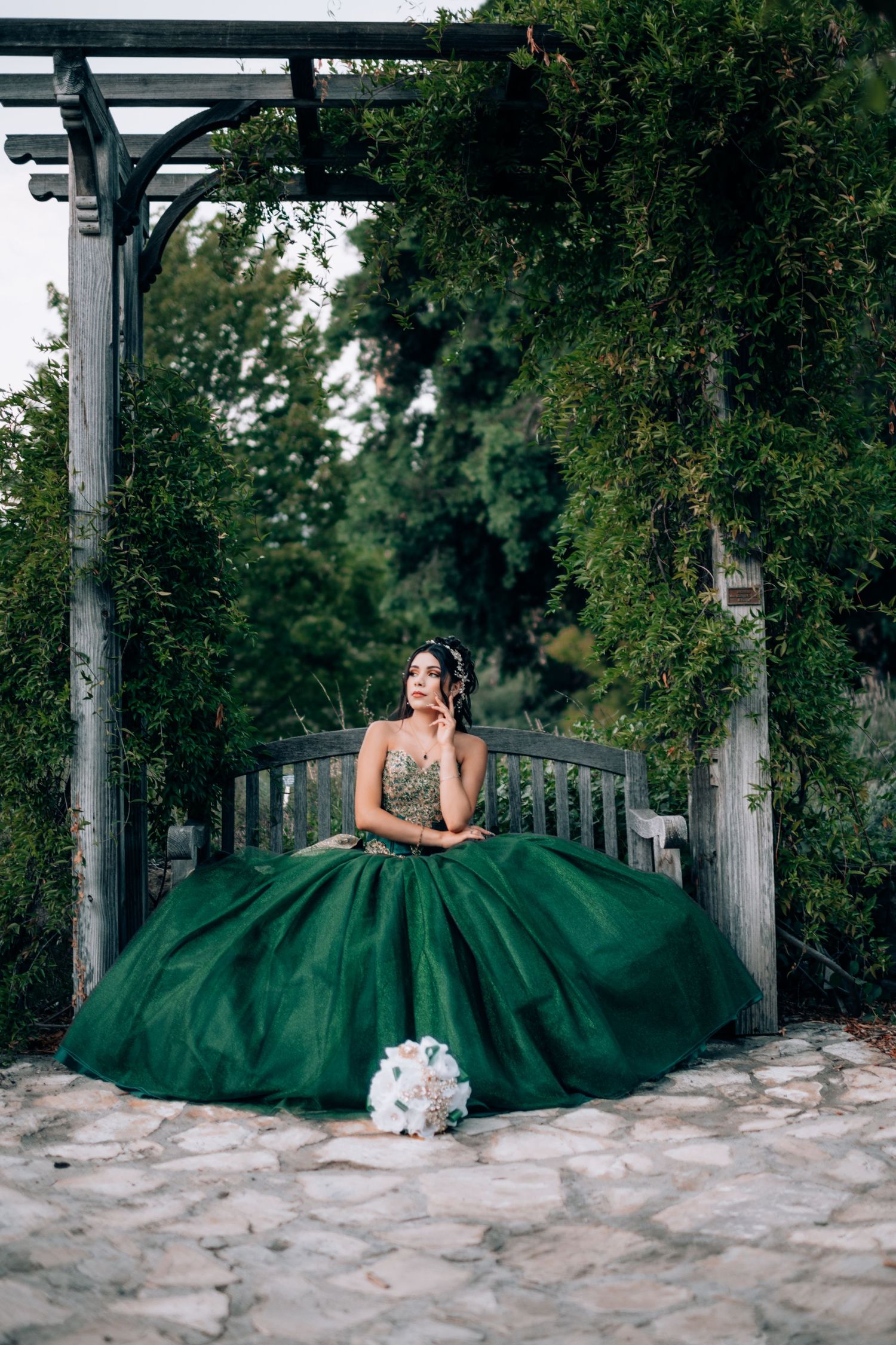 Elegant emerald green quinceañera gown with full tulle skirt photographed in romantic garden setting with white bouquet.