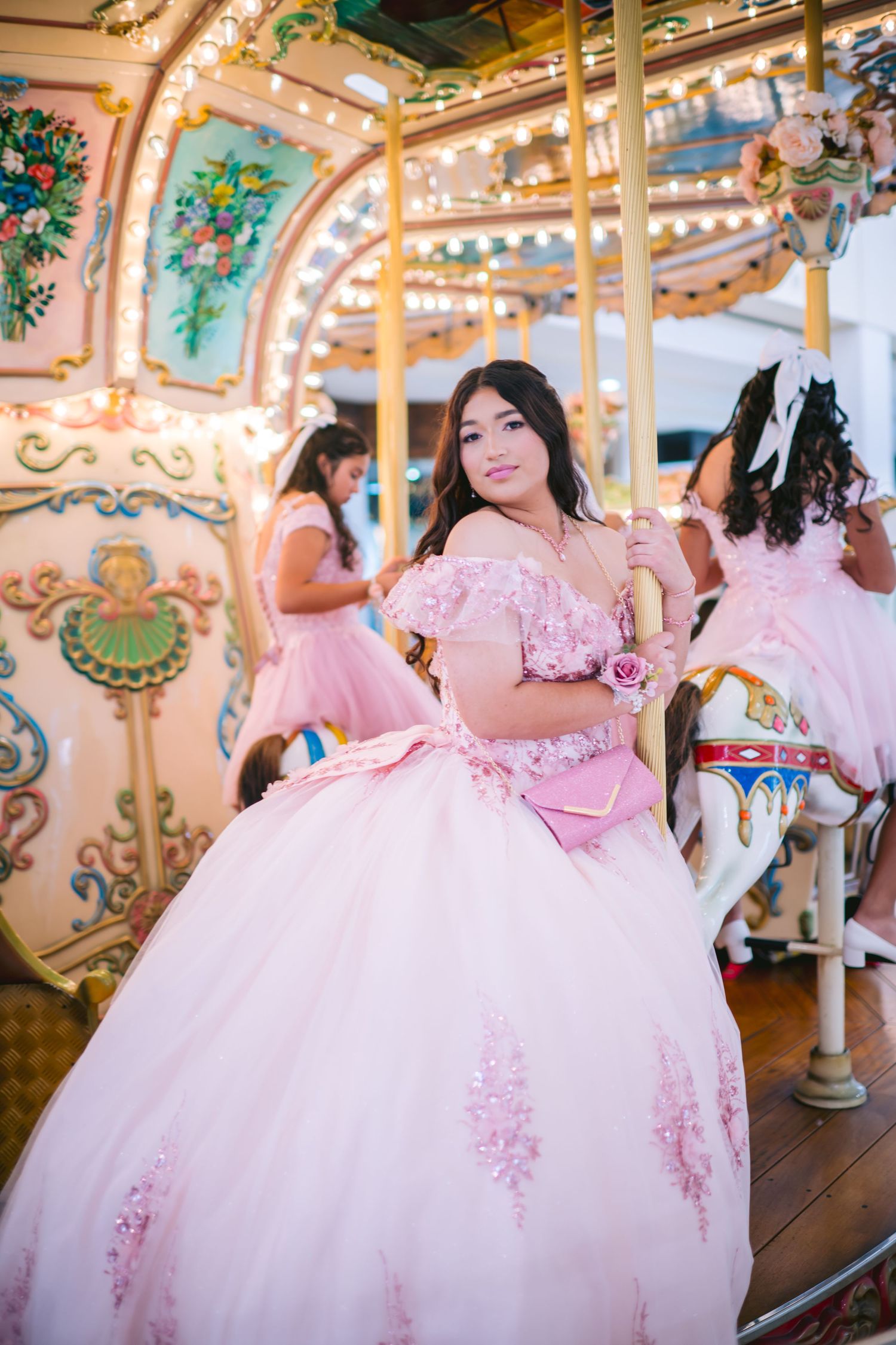 A person in a pink ball gown poses on an ornate vintage carousel with decorative horses and floral details.