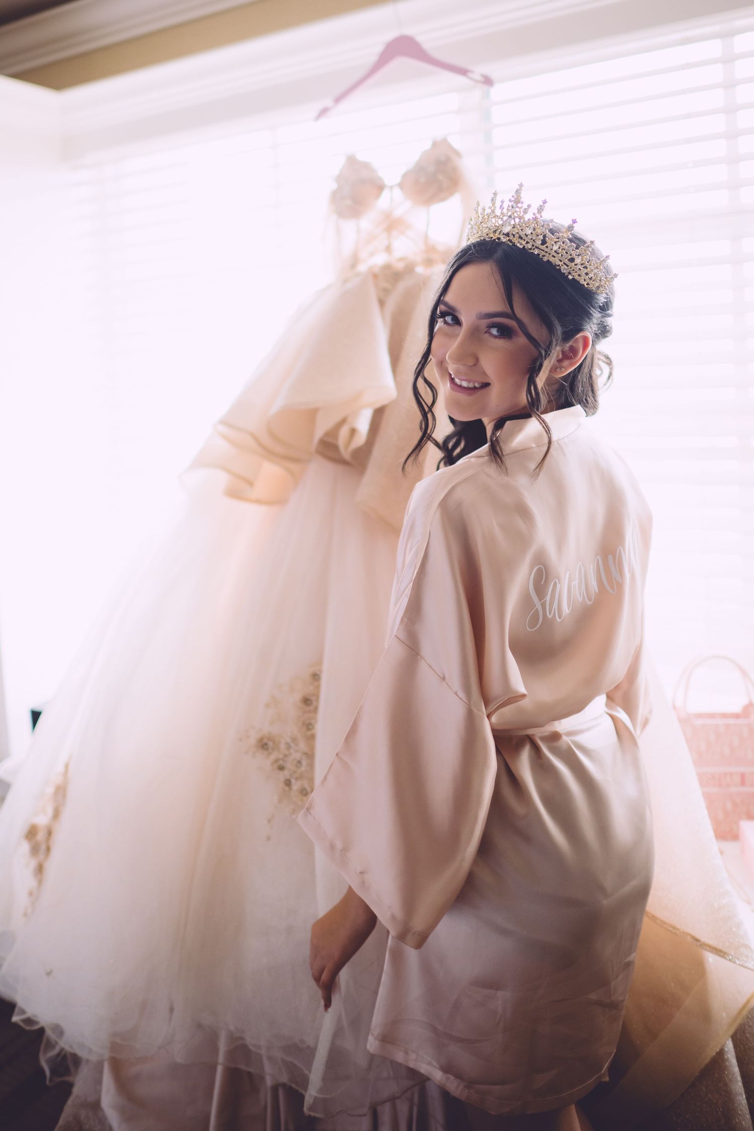 A bride in a white robe and tiara poses near a hanging wedding dress during wedding preparations.