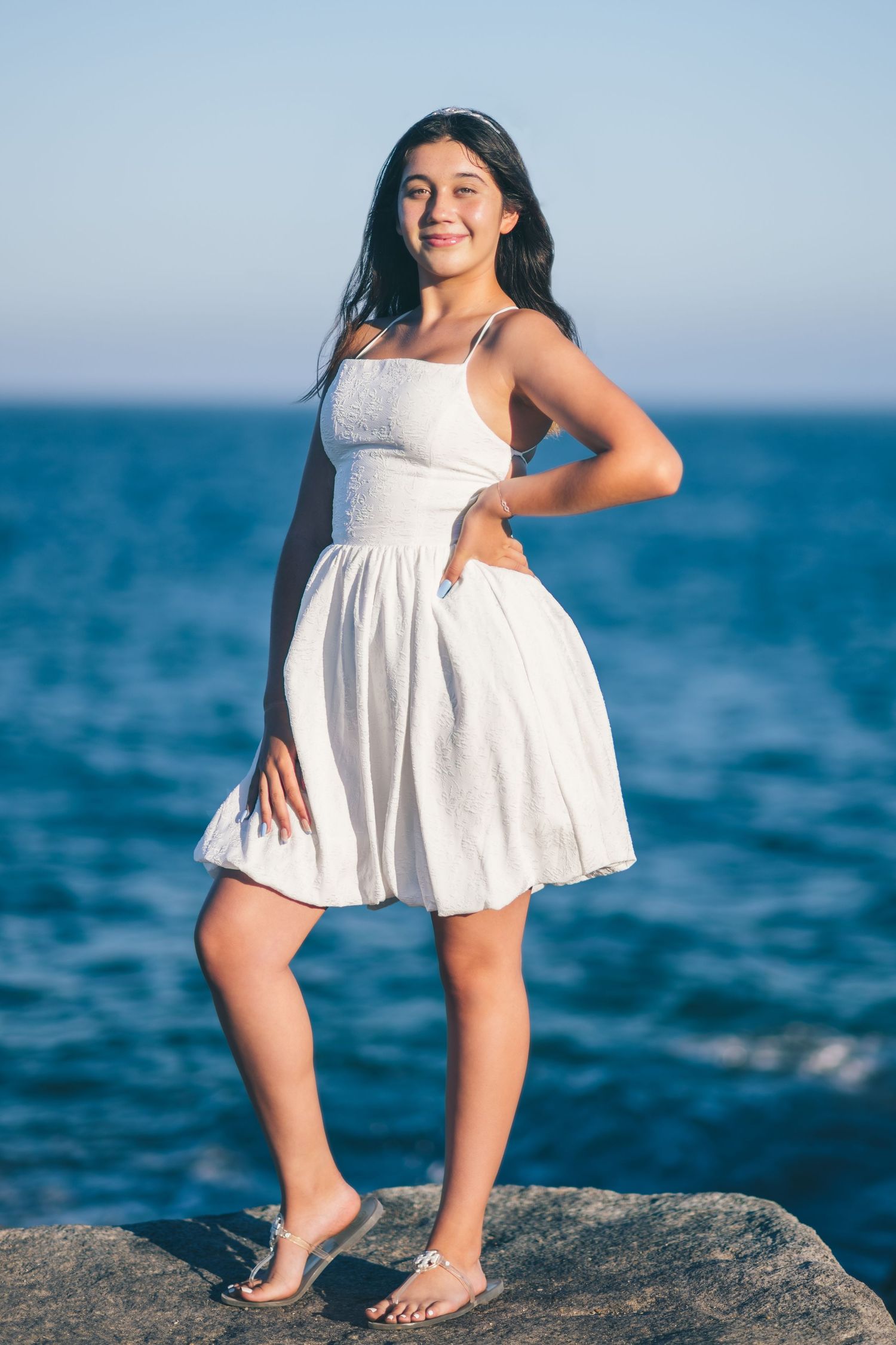 A person in a flowy white summer dress poses confidently on rocky coastal shoreline with blue ocean waves in background.