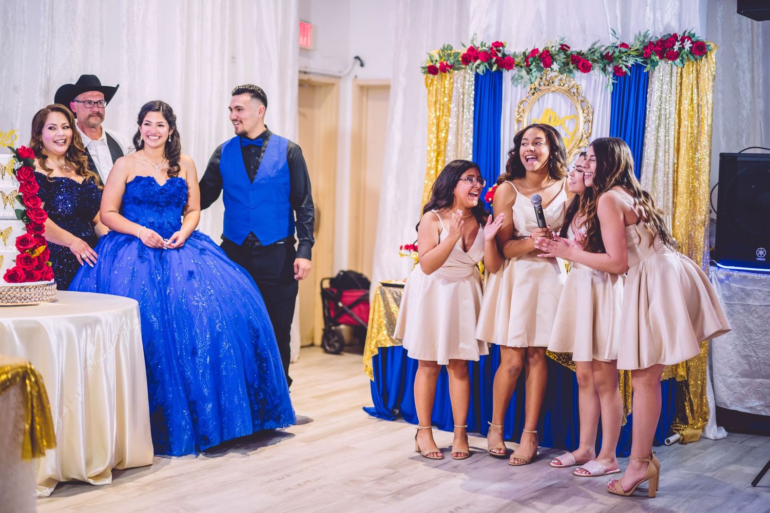 A celebratory event with people in formal wear, featuring royal blue and pink dresses against an elegant white backdrop.