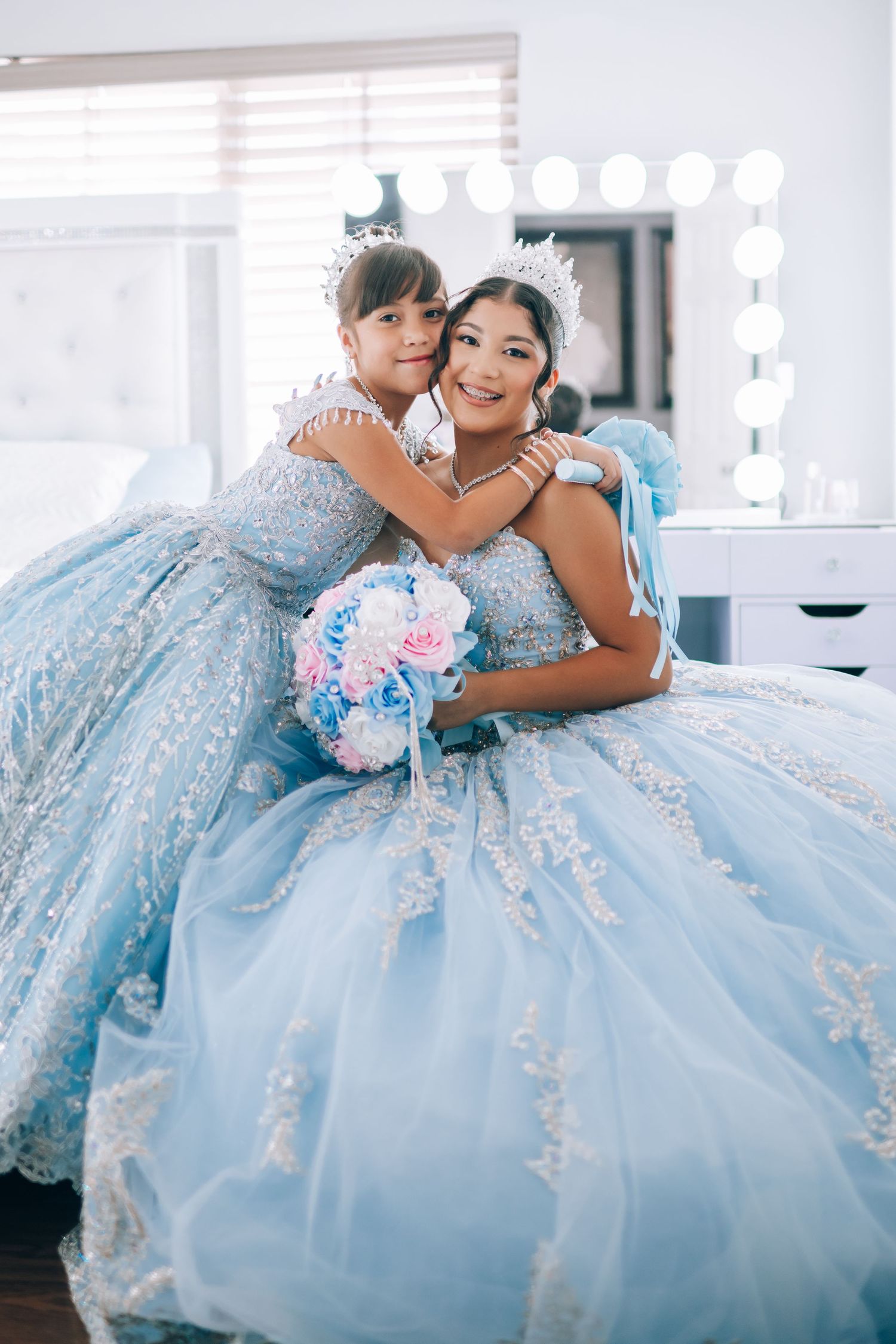 Two people wearing matching light blue princess ball gowns with lace details sit together holding pink and blue bouquet.