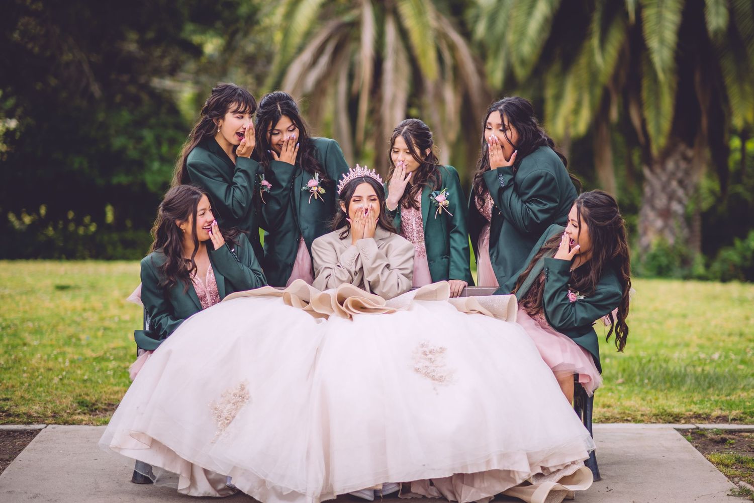 A bride in a white ball gown sits surrounded by bridesmaids in green dresses on grass with palm trees in background.