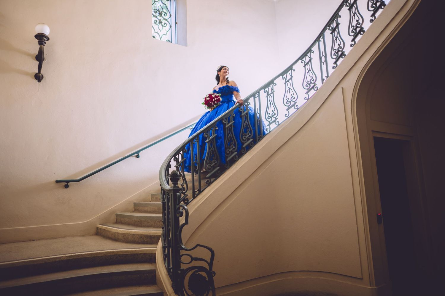 A person in a bright blue formal gown poses elegantly on a curved staircase with ornate iron railings.