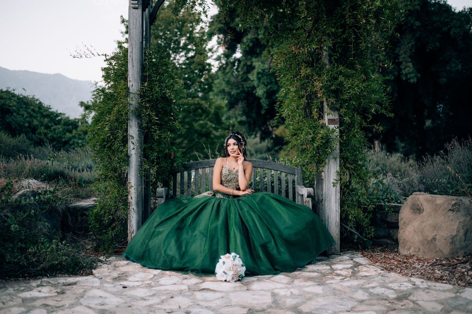 Young woman in an elegant emerald green ball gown quinceañera dress poses gracefully in a lush garden setting.