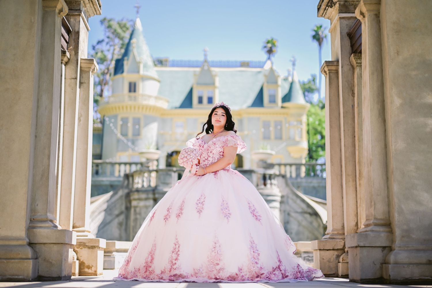 A pink ballgown flows elegantly across a stone balcony with ornate architecture and castle towers in the background.