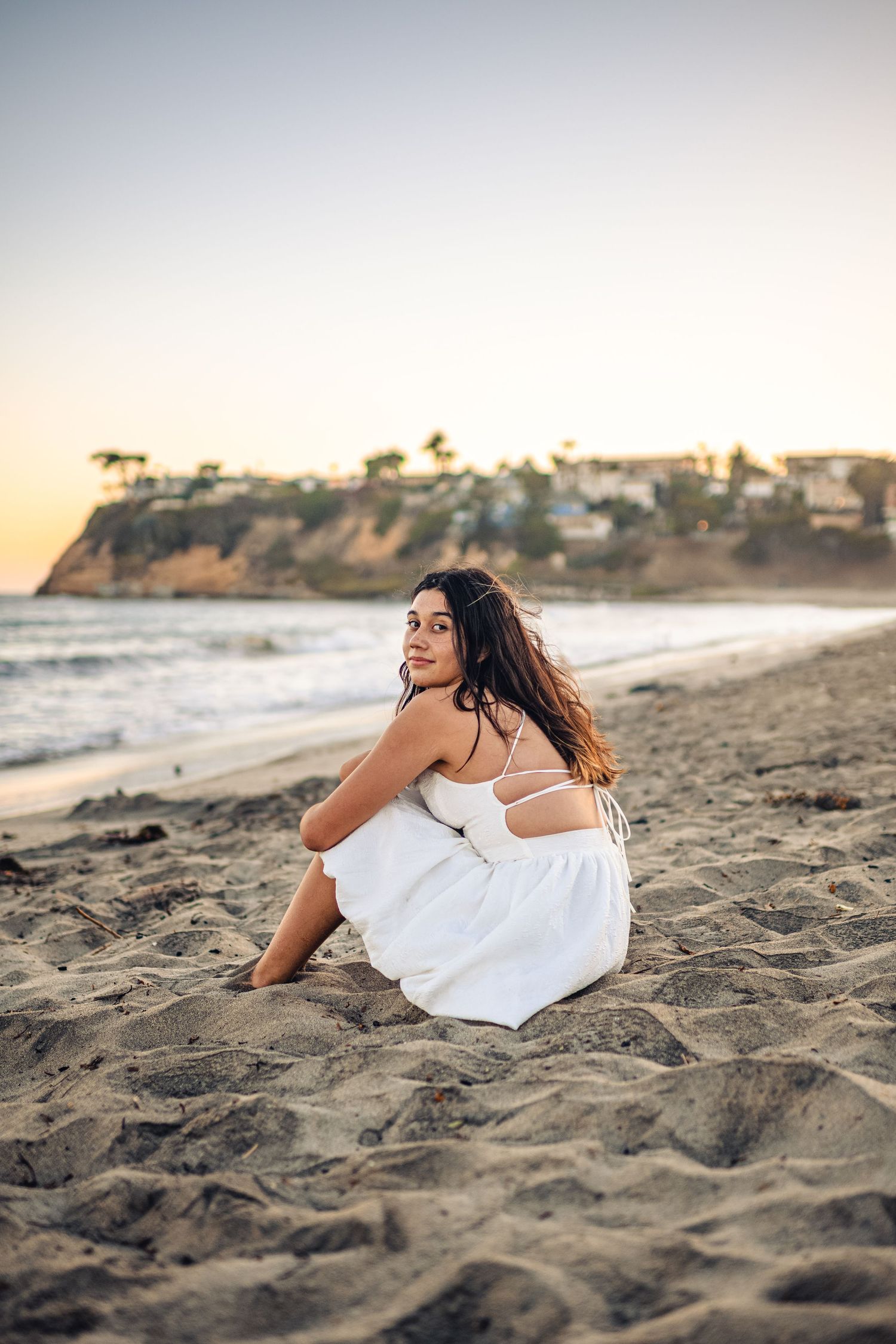 Person in white dress sits on sandy beach at sunset with coastal cliffs and ocean waves in background.