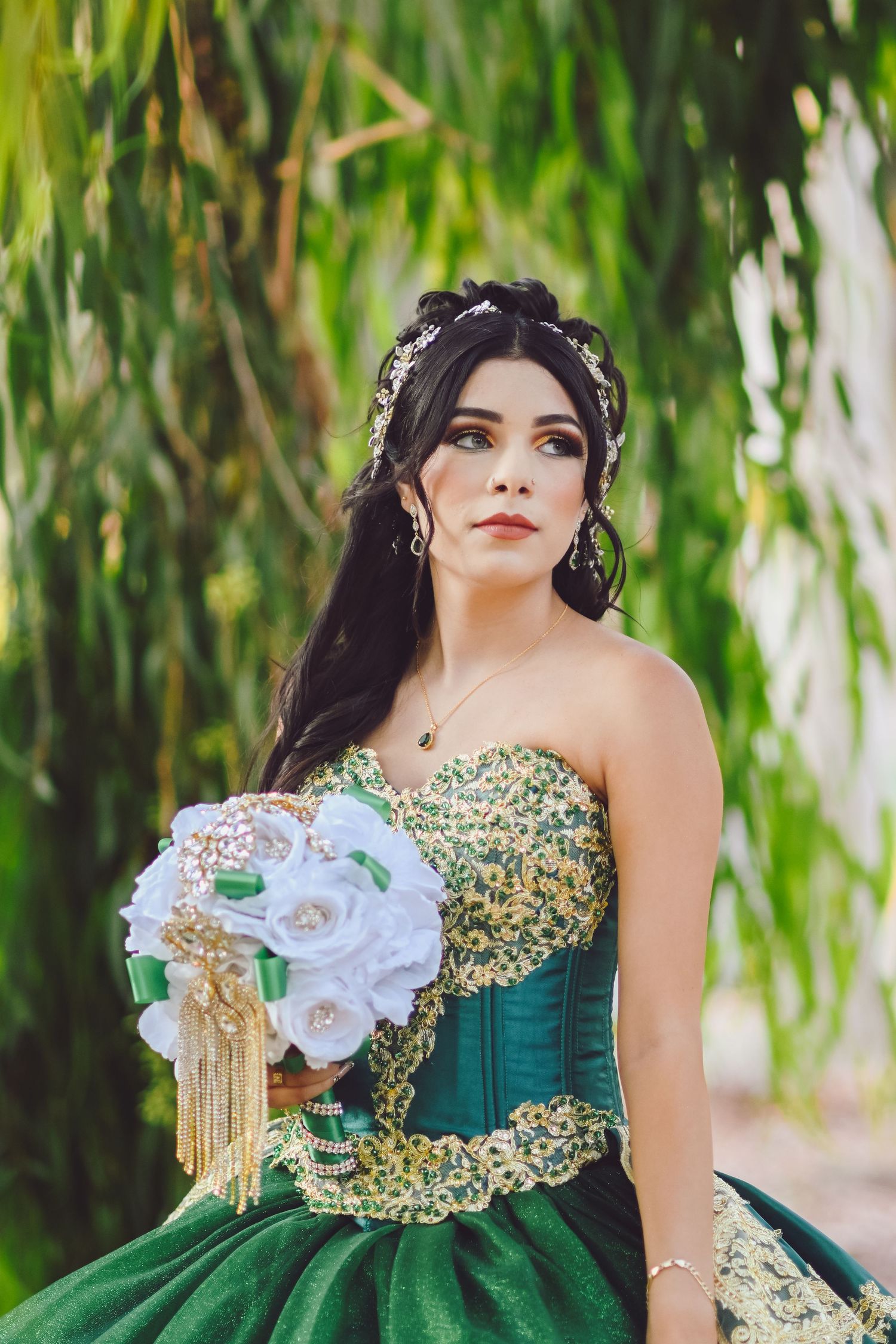 Emerald ball gown with gold beading details and white floral bouquet against willow tree backdrop.