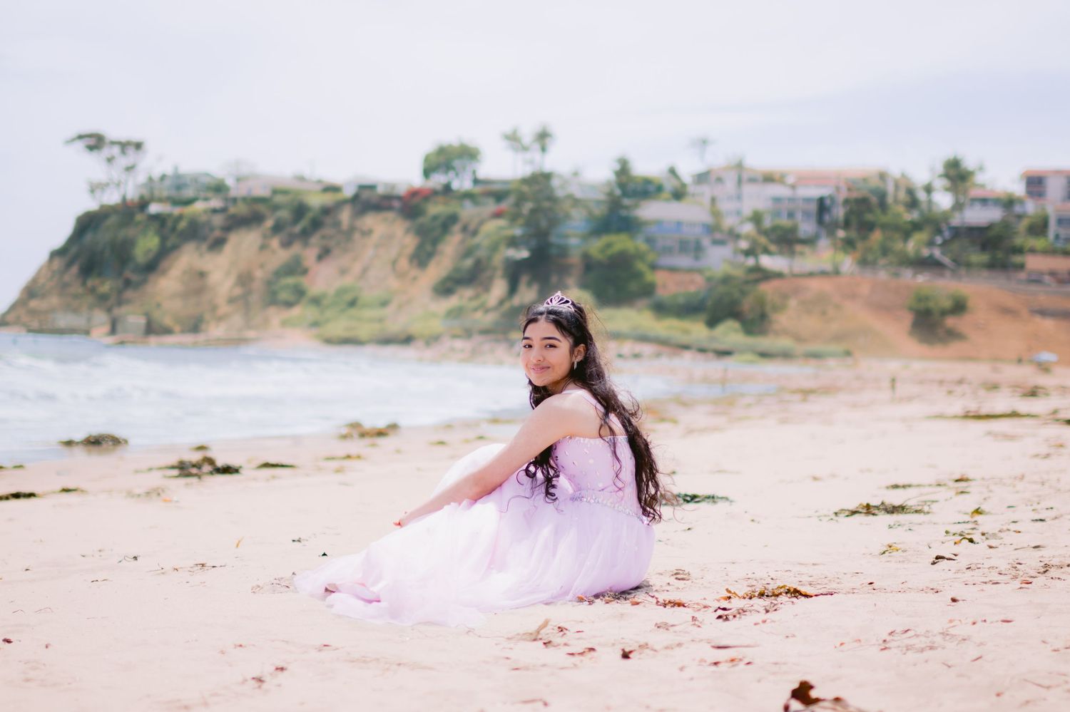 A young girl in a lavender tulle dress sits on a sandy beach, smiling over her shoulder with a coastal bluff behind her.