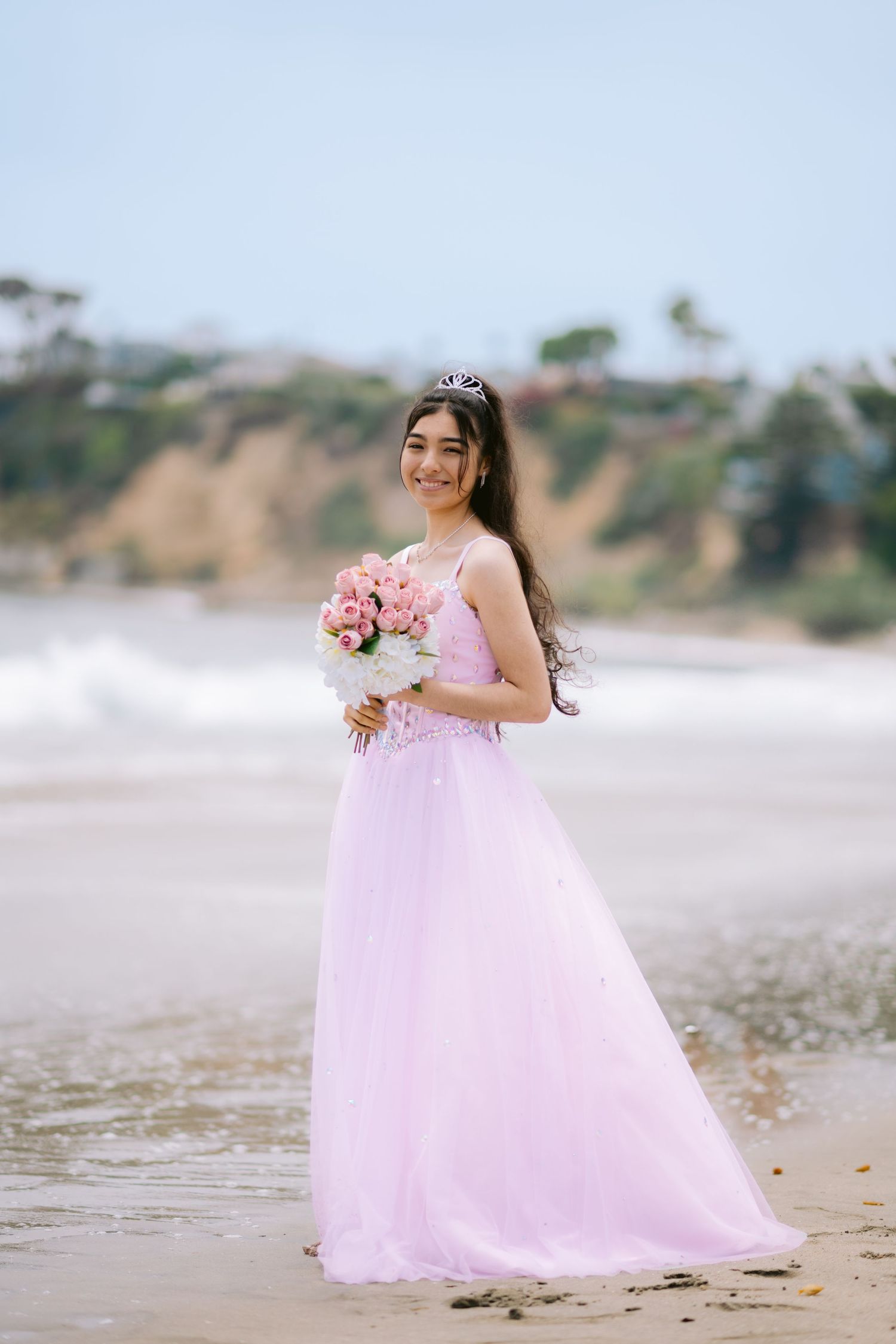A person in a flowing pink gown and tiara holds a bouquet while standing on a beach with cliffs in the background.